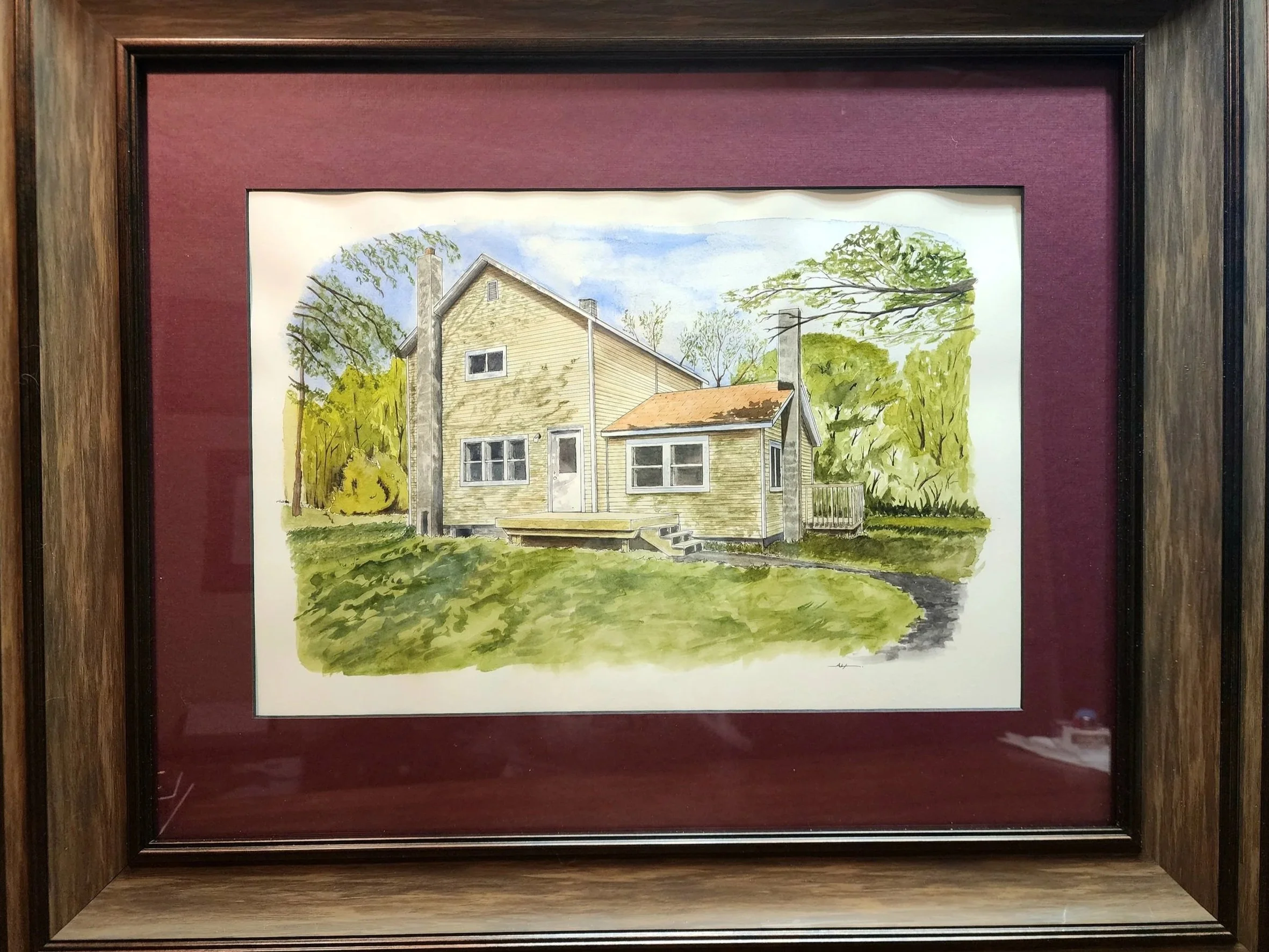 Framed watercolor painting of a tan house with a porch, surrounded by trees, under a blue sky.