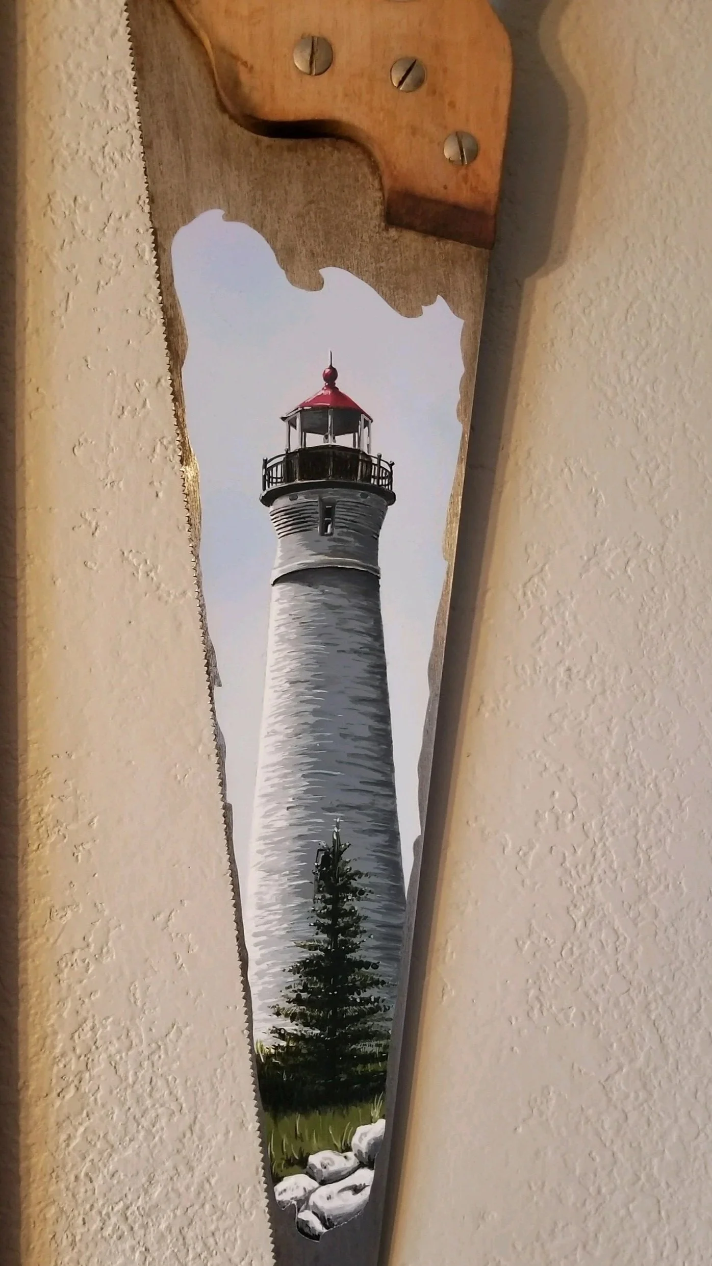 Painted lighthouse on an old handsaw with rocks and trees at the base.