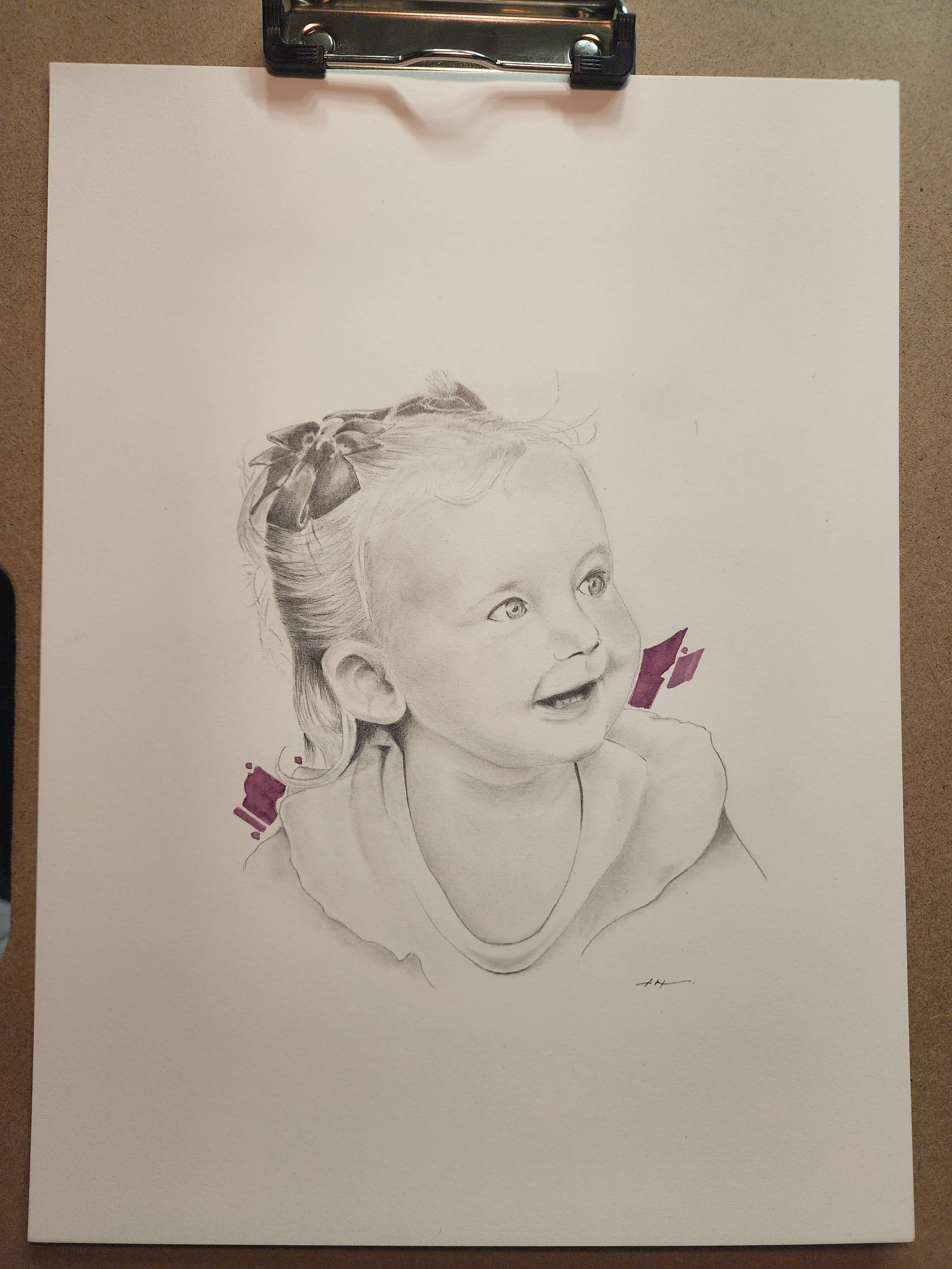 A detailed graphite pencil drawing of a young child's face on a white sheet of paper, with some shaded areas and purple paint strokes, held in place with a black clipboard on a brown surface.