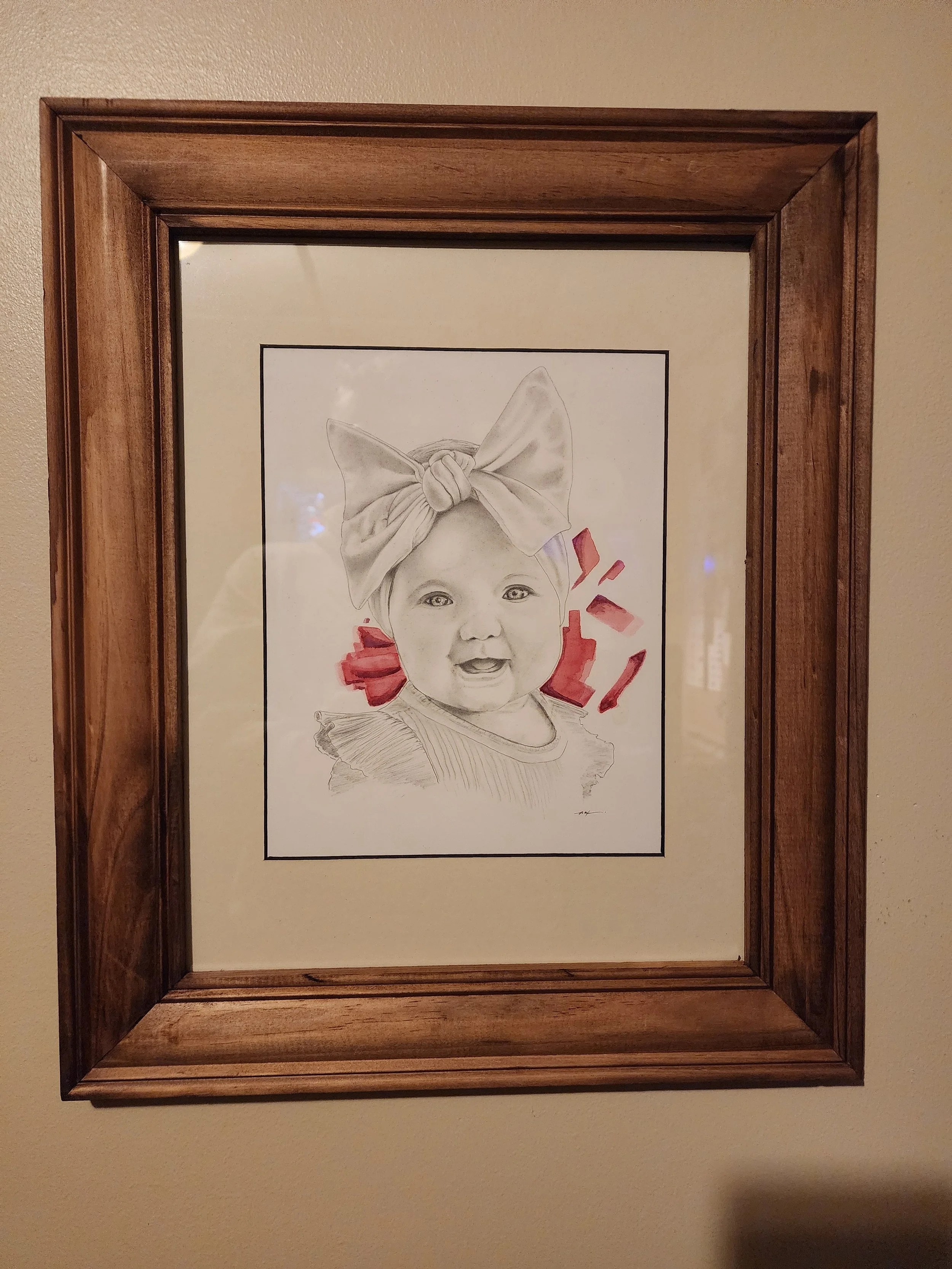 A framed drawing of a child's face with a large bow on her head and pink accents around her neck.