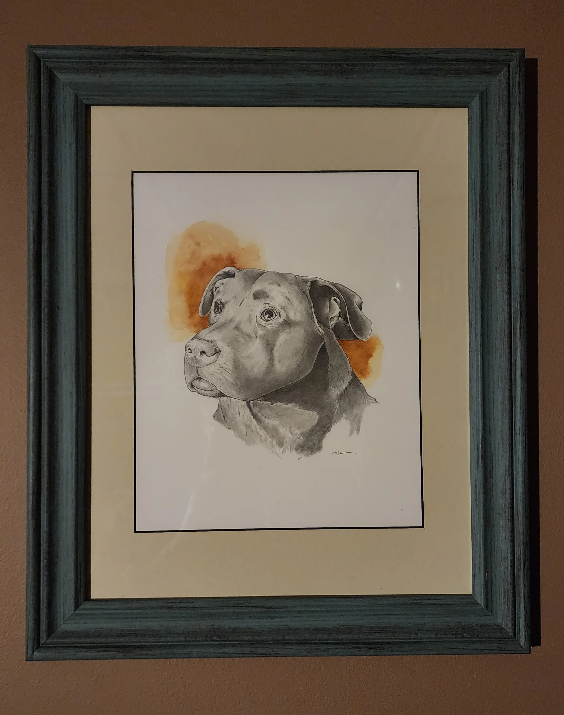 A framed watercolor portrait of a dog, possibly a Labrador Retriever, depicted in a realistic style with a yellowish-orange watercolor background, hung on a brown wall.