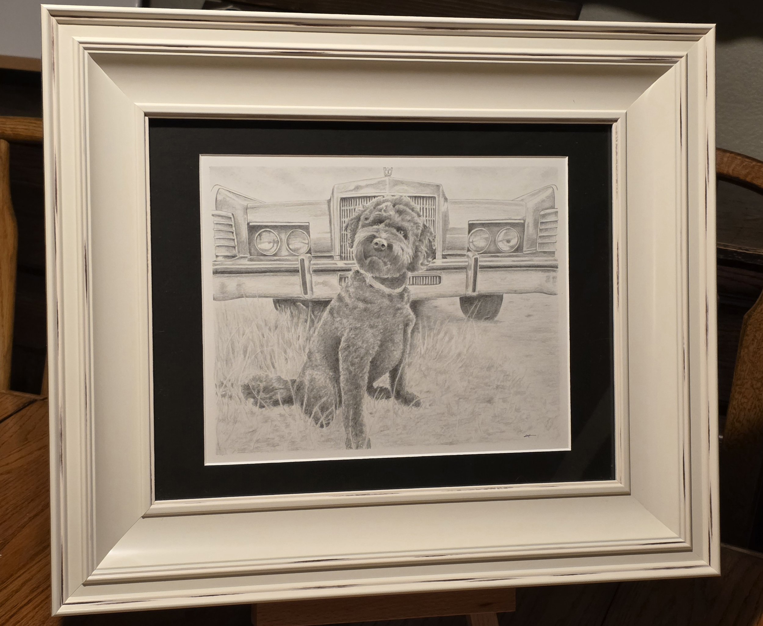 A framed black and white pencil drawing of a dog sitting in front of an old car.