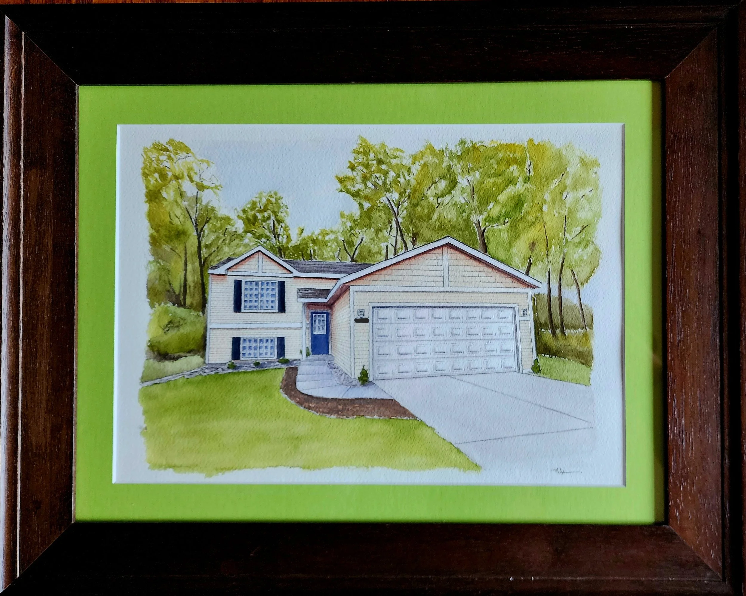 Framed watercolor painting of a house with a garage, surrounded by trees, with a bright green mat and a dark wooden frame.