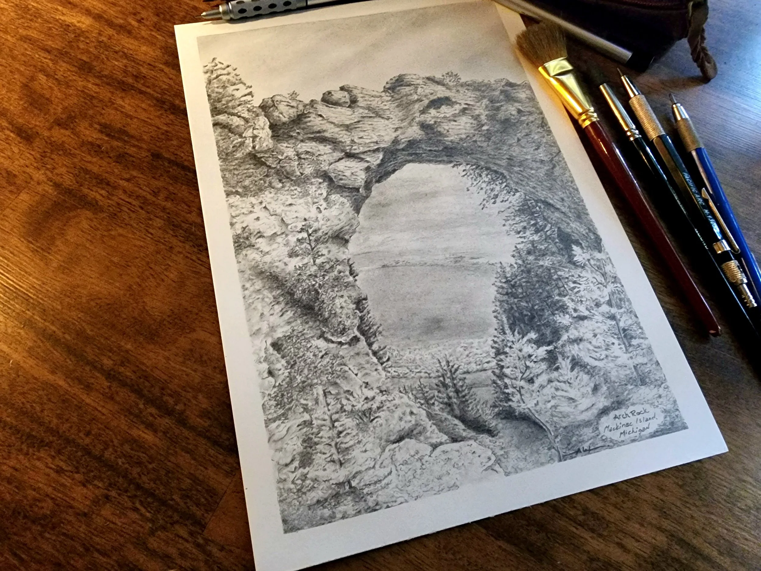 A black and white pencil drawing of a natural rock formation with an arch, trees, and a landscape, titled 'Arch Rock, Mackinac Island, Michigan'