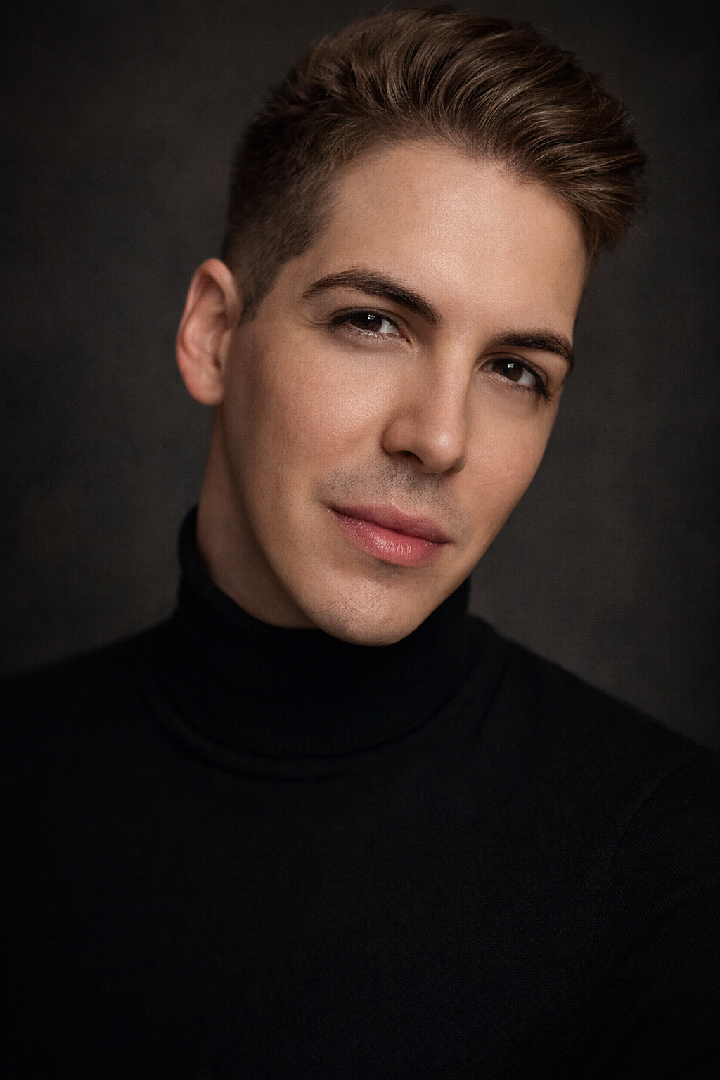 A portrait of a young man with short, styled hair wearing a black turtleneck against a dark background.