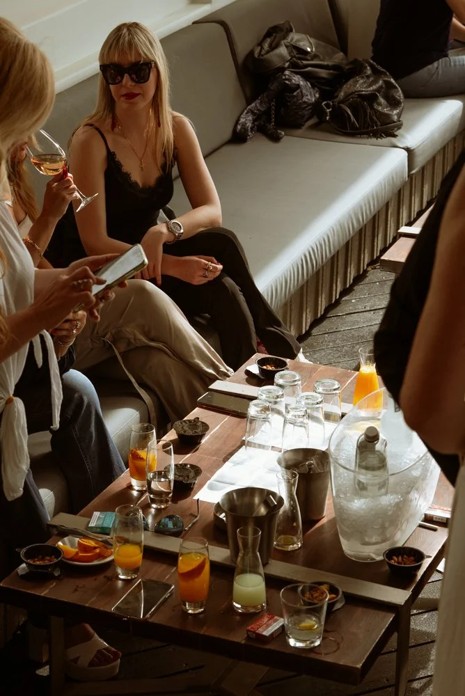 A woman with long blonde hair, wearing sunglasses and a black tank top, sitting on a gray bench, holding a glass of wine, and looking at her phone. There are drinks and snacks on the table in front of her, and other people are partially visible aroun