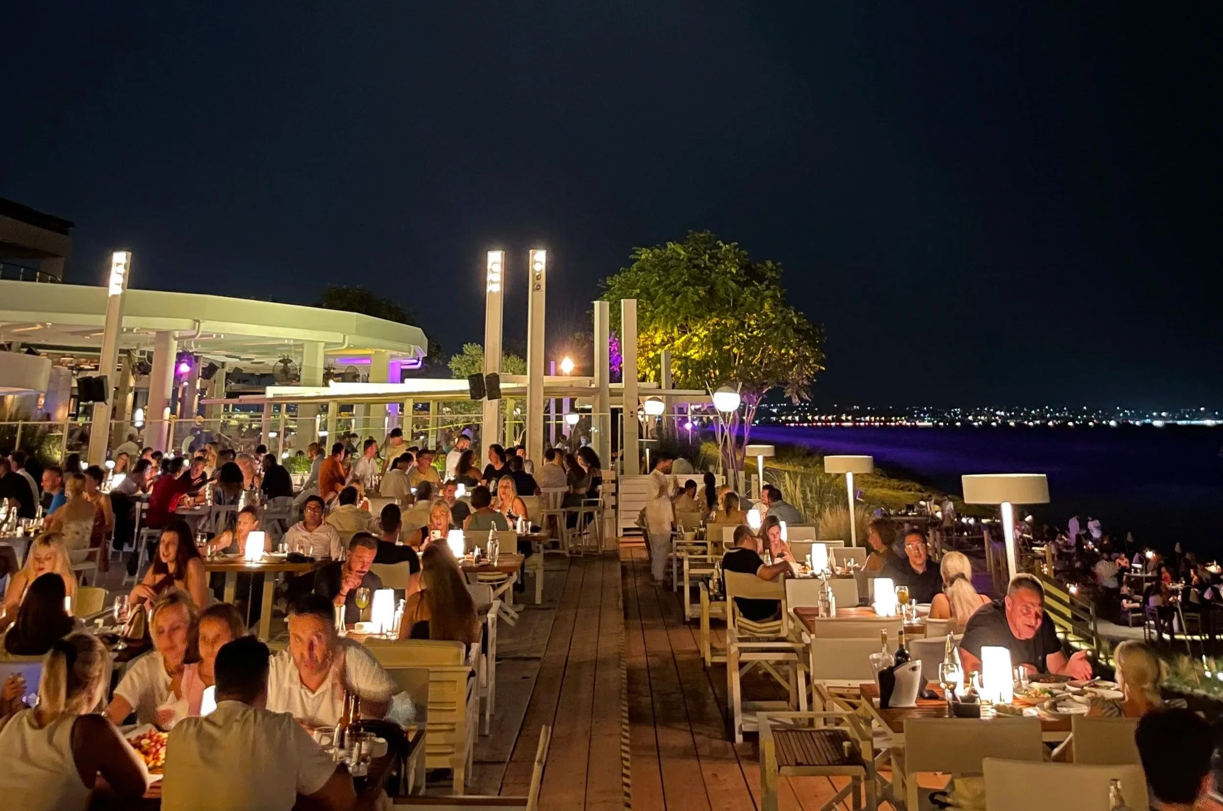 Nighttime scene of a bustling outdoor restaurant by the water, with many tables filled with people dining under warm lighting, and city lights in the distance.