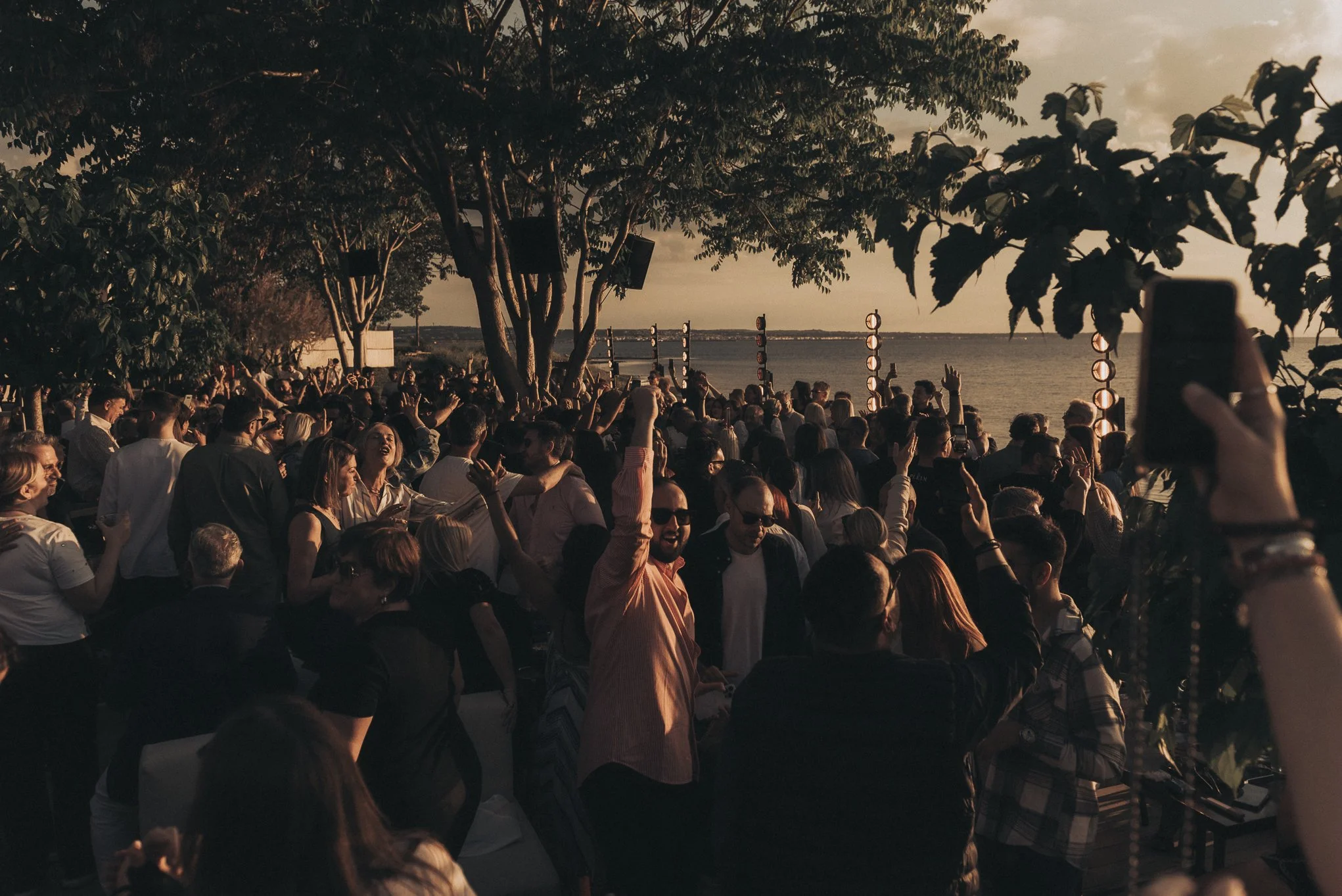 People gathered outdoors by the water at sunset, enjoying a social event with some raising their hands and taking pictures.