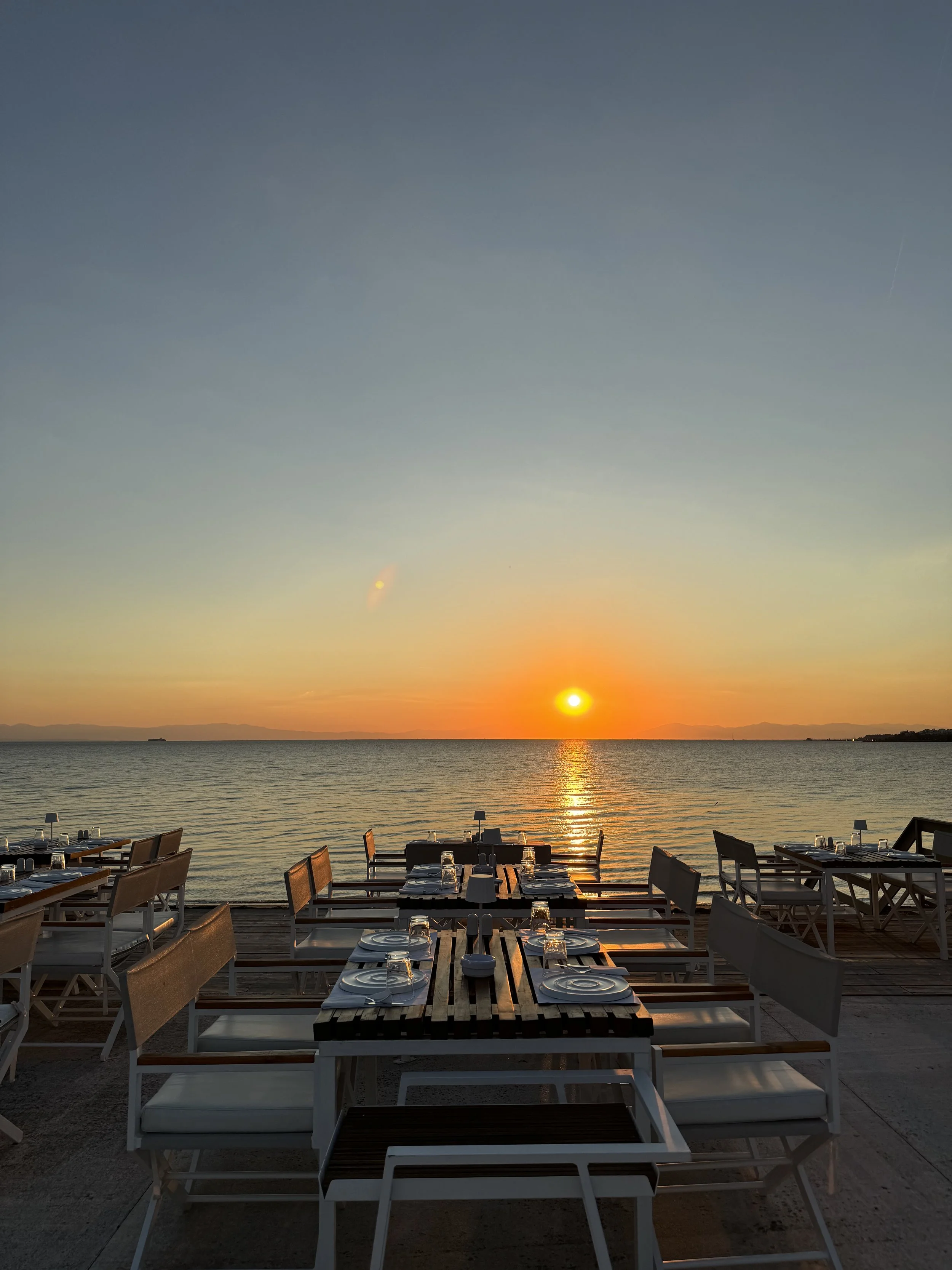 Outdoor dining tables with plates and glasses set up on a deck facing the ocean at sunset, with the sun near the horizon and a calm sea.