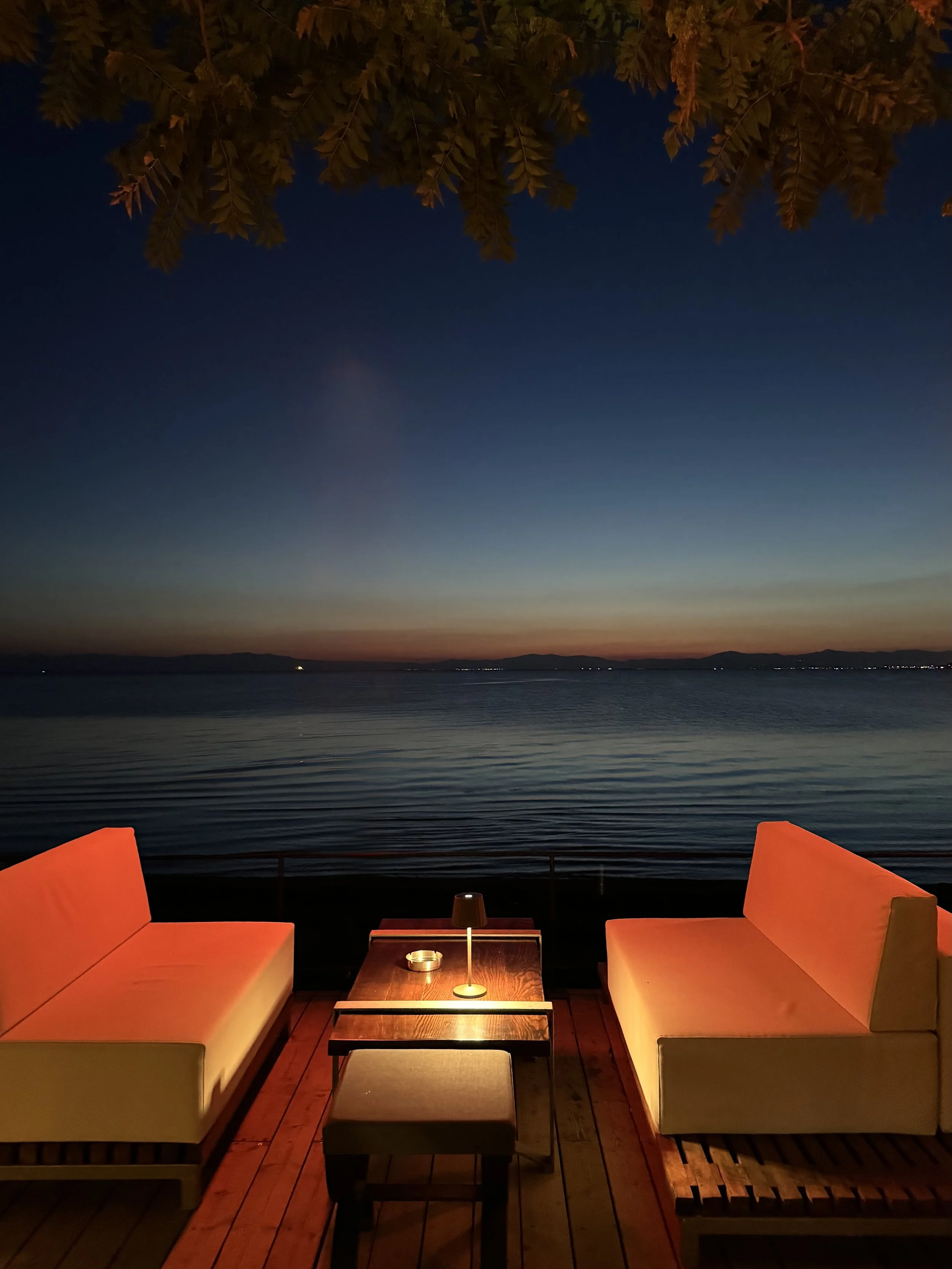 A lakeside outdoor seating area at dusk with two modern white sofas, a wooden table with a small lamp and candle, and a tree with leaves overhead, overlooking a calm lake under a darkening sky.