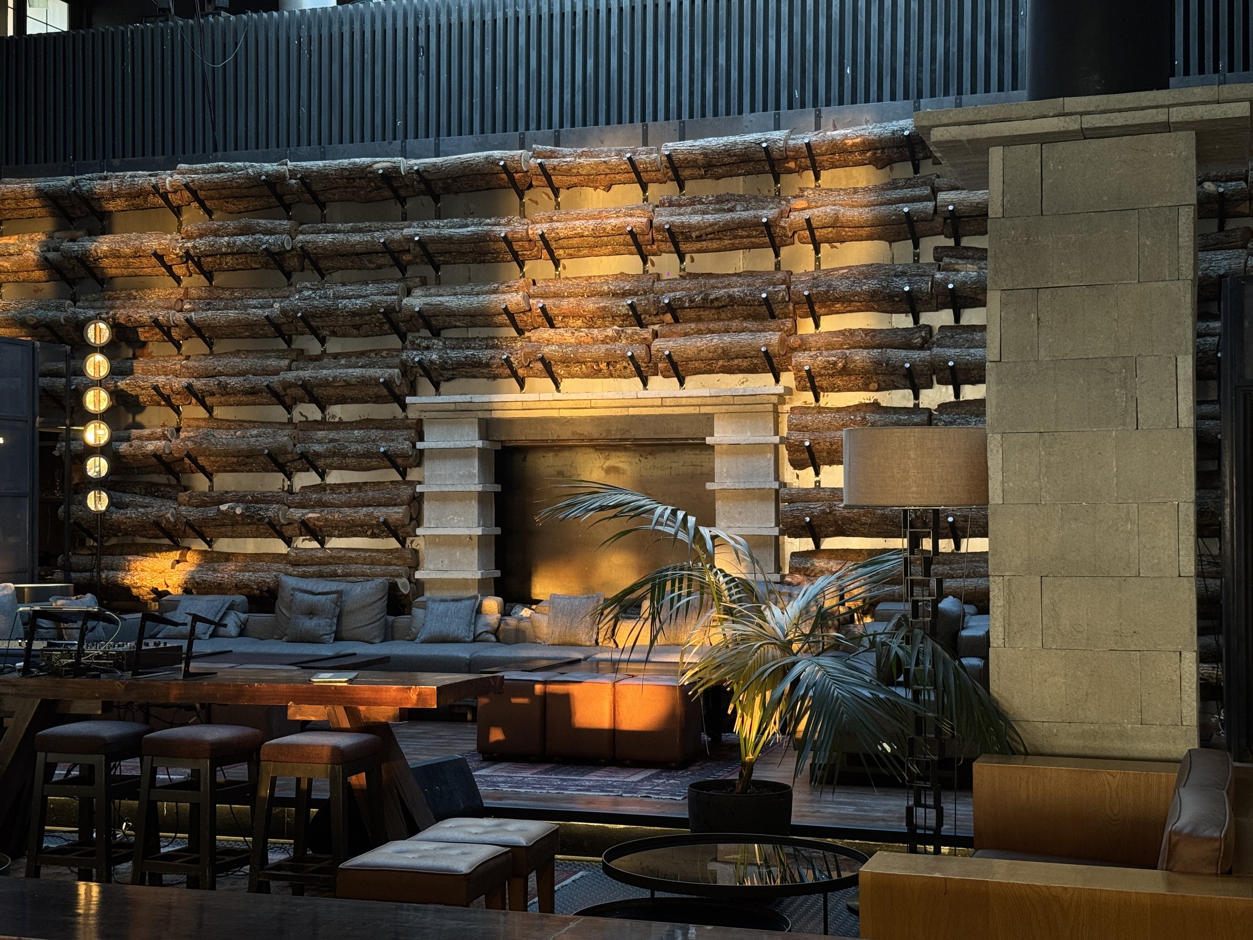 Indoor lounge area with a stone fireplace, a sofa with multiple cushions, potted plants, wooden furniture, and a lit wall with stacked logs decor.