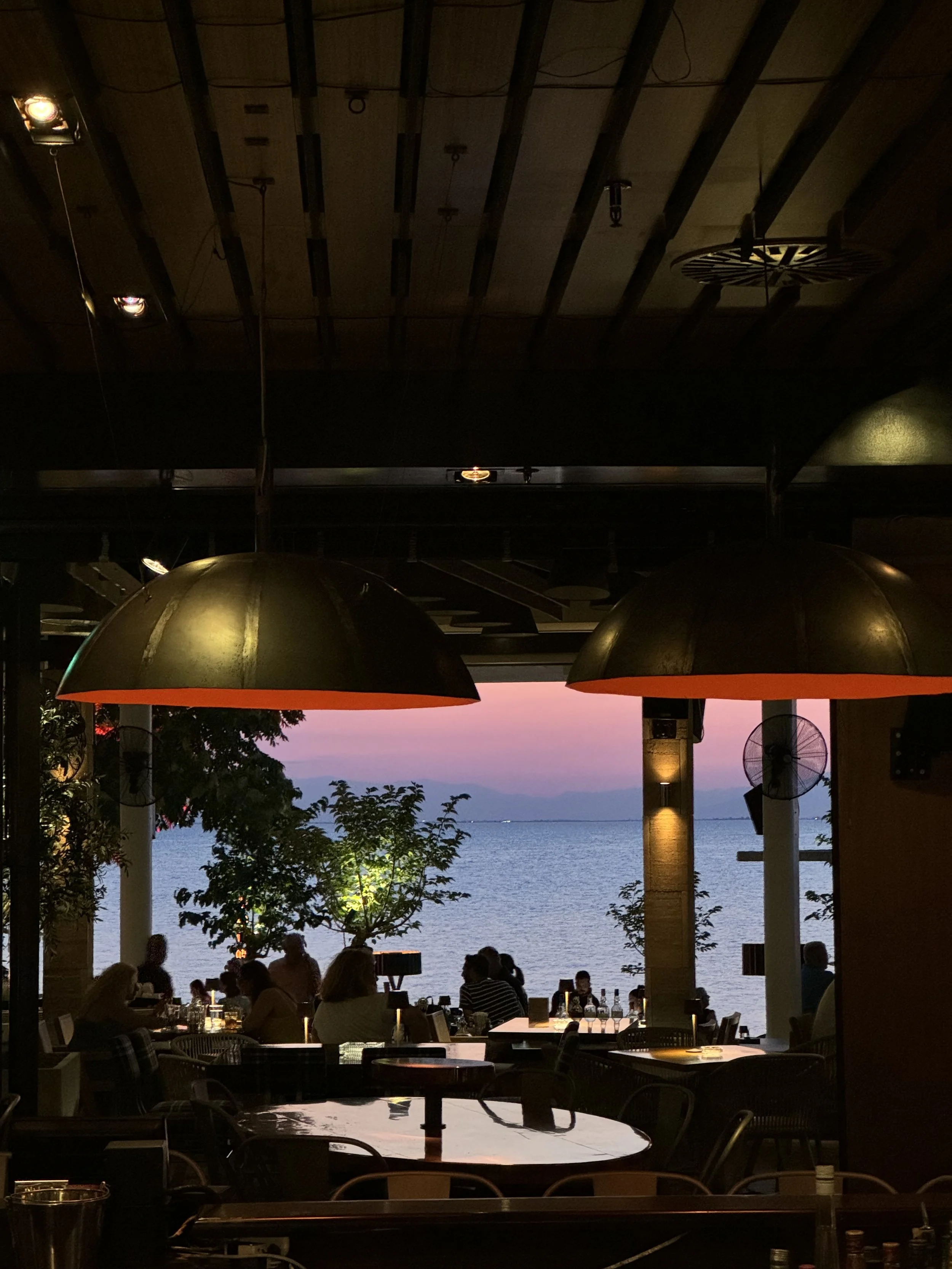 Dining area in a restaurant with large hanging lamps, overlooking a body of water during sunset, with several people seated at tables and trees outside.