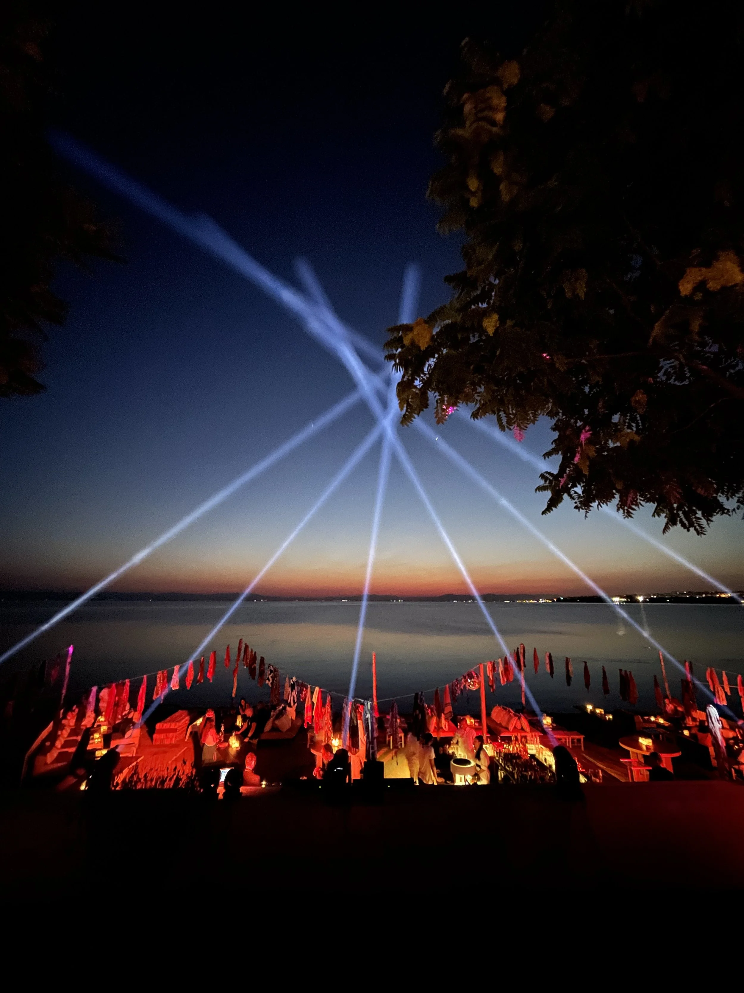 Nighttime scene at a waterfront with laser light show, a large tree partially seen on the right, and tents or booths illuminated in red along the shoreline.