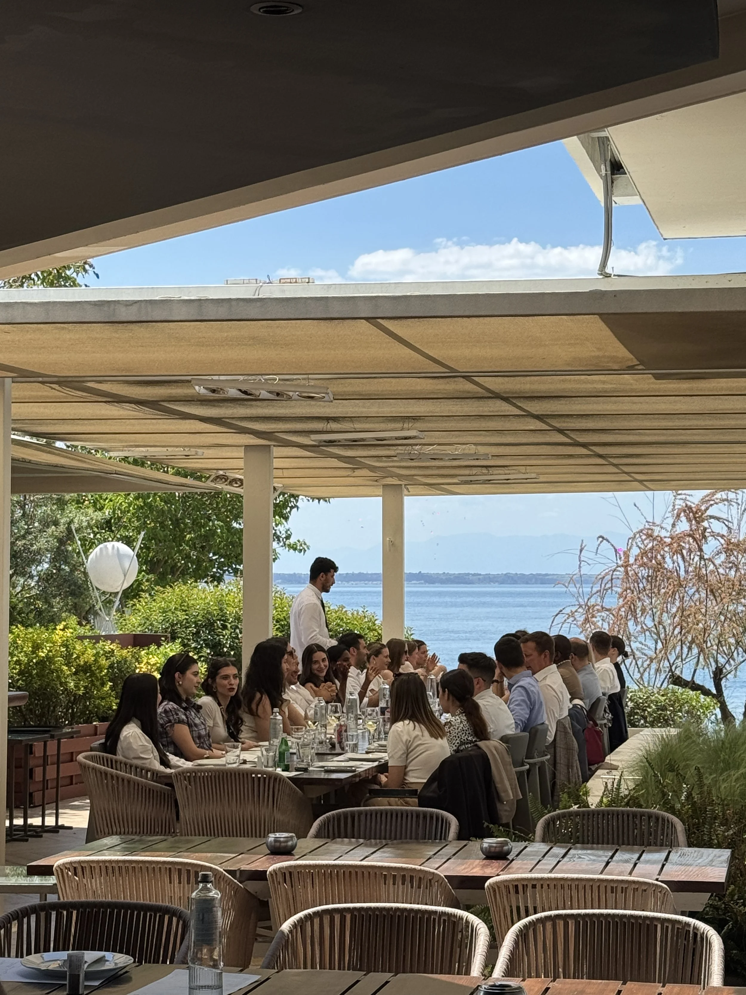 People dining at an outdoor restaurant with a view of the water and blue sky.