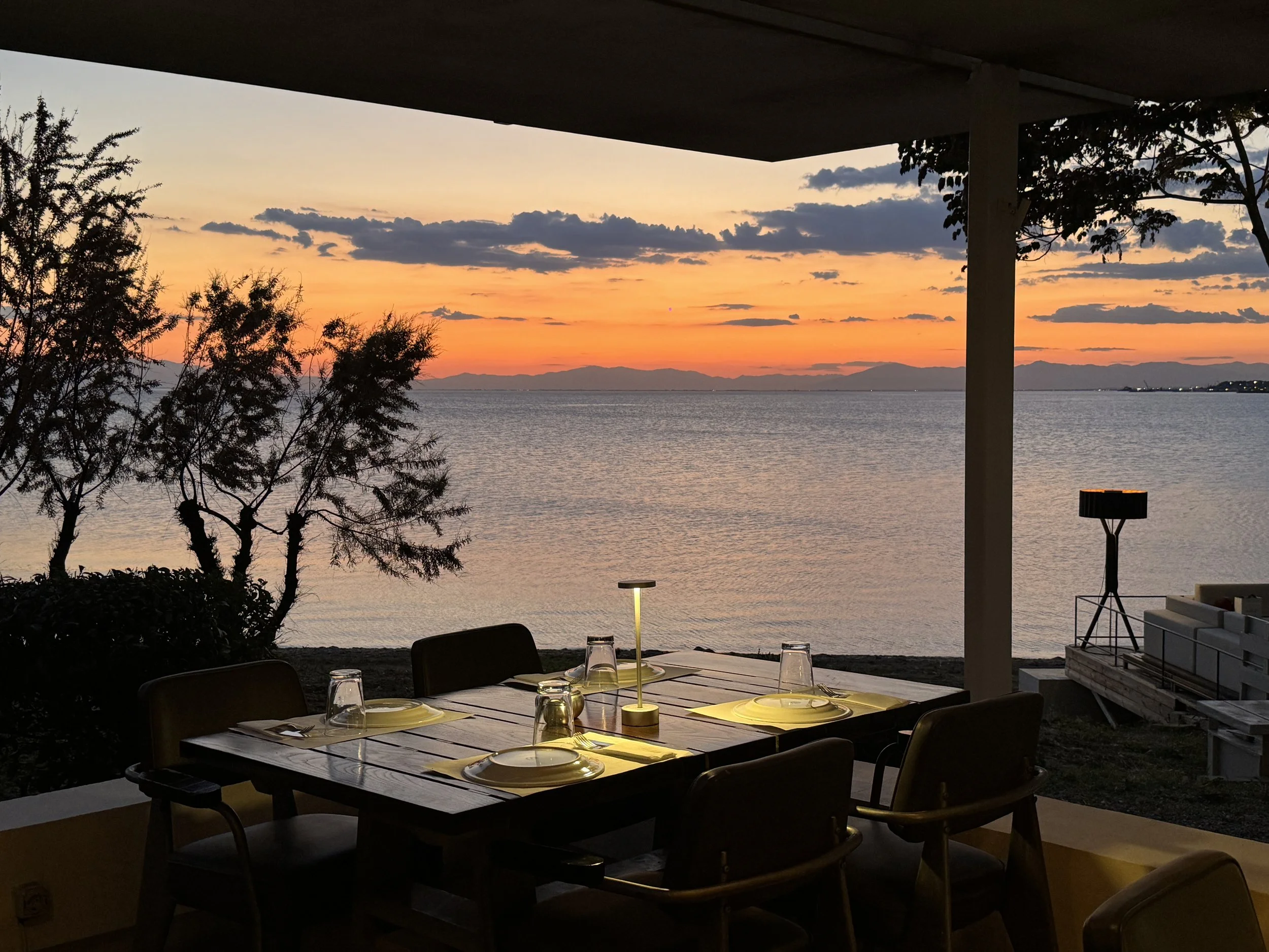 A dining table set for four with plates, glasses, and cutlery, under a covered patio overlooking a body of water at sunset with orange, pink, and purple sky, trees, and distant mountains.
