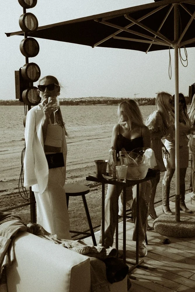 People gathered on a beachside bar or lounge area during sunset, with a woman in sunglasses taking a selfie, others socializing near the water, under a large umbrella.