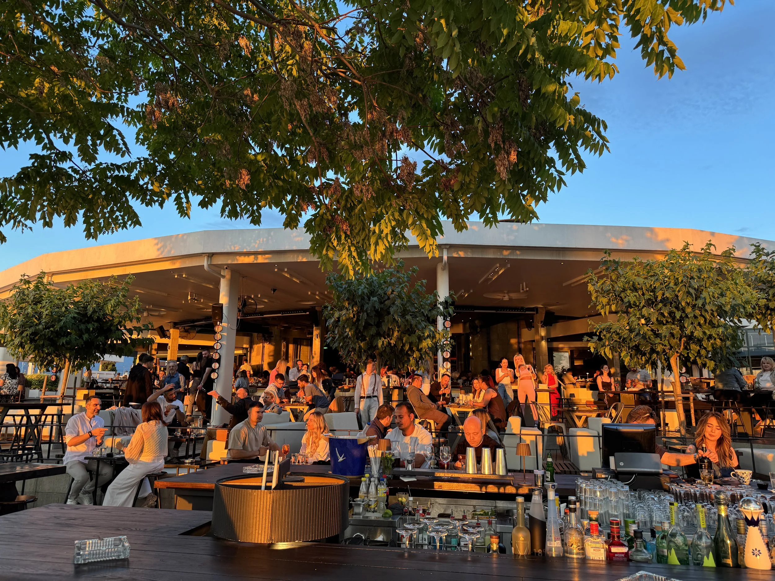 People enjoying drinks and socializing at an outdoor rooftop bar or restaurant during sunset, with trees and a modern building in the background.