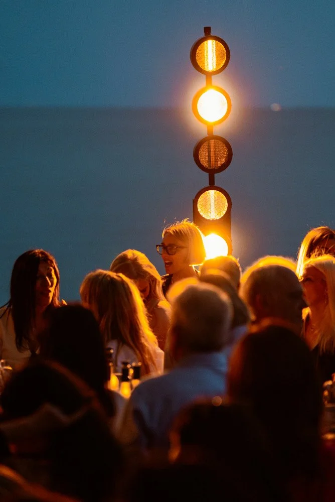 A group of people gathered outdoors at sunset, illuminated by a traffic light with glowing yellow and red lights, with a clear sky in the background.