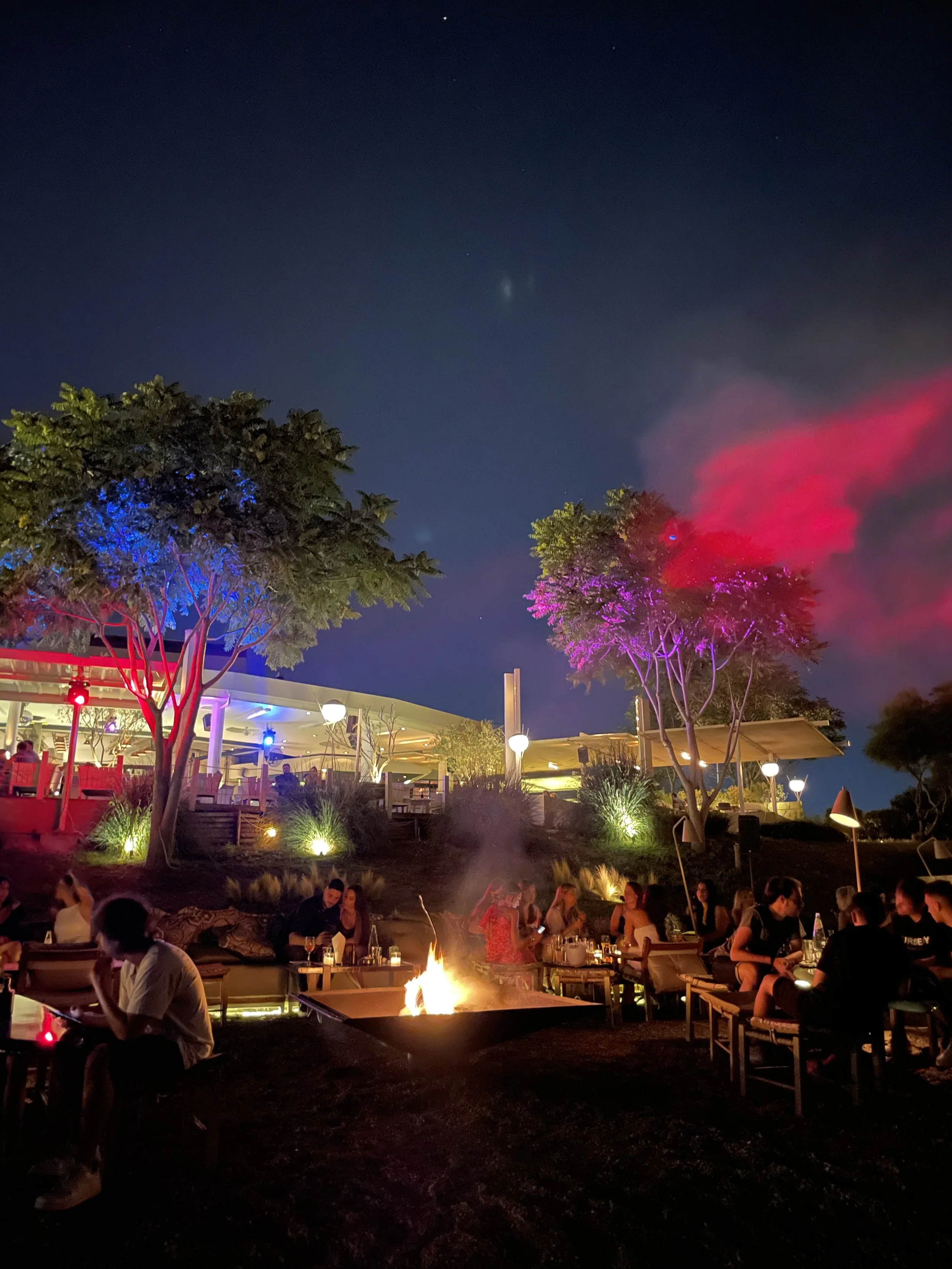 Night scene of an outdoor restaurant or bar with people sitting around tables near a fire pit. The area is decorated with colorful lighting, trees, and illuminated lamps. The sky is dark with visible stars.