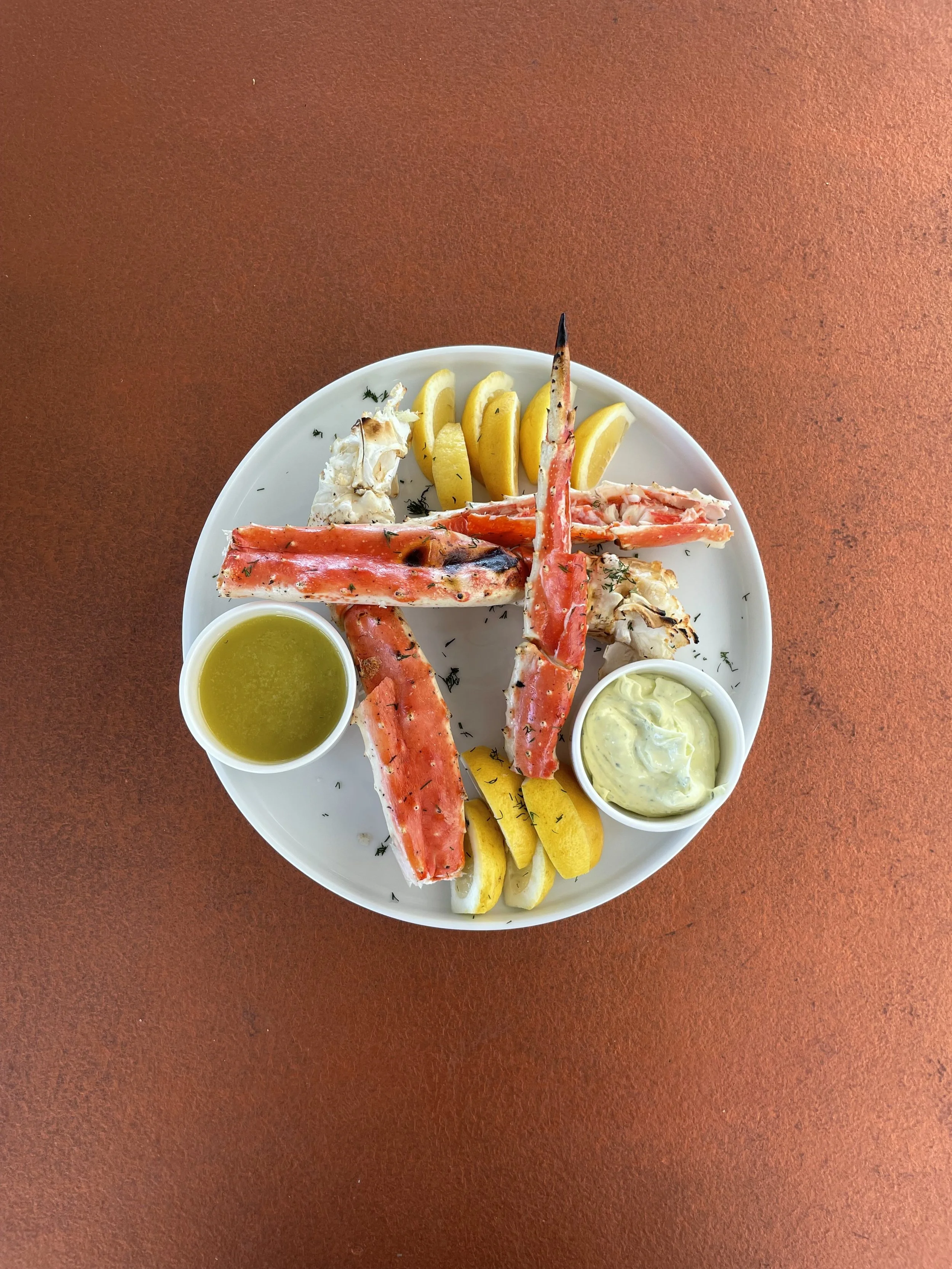Plate with cooked crab legs, lemon wedges, a small cup of melted butter, and a small cup of dipping sauce on a reddish table.