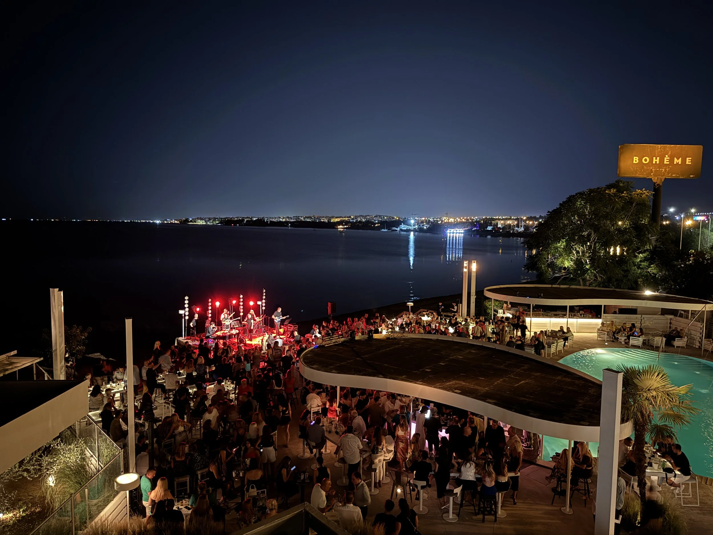 Nighttime scene at Boheme, an outdoor restaurant and bar by a waterfront, with a live band performing on stage, patrons sitting around tables, and city lights reflecting on the water.