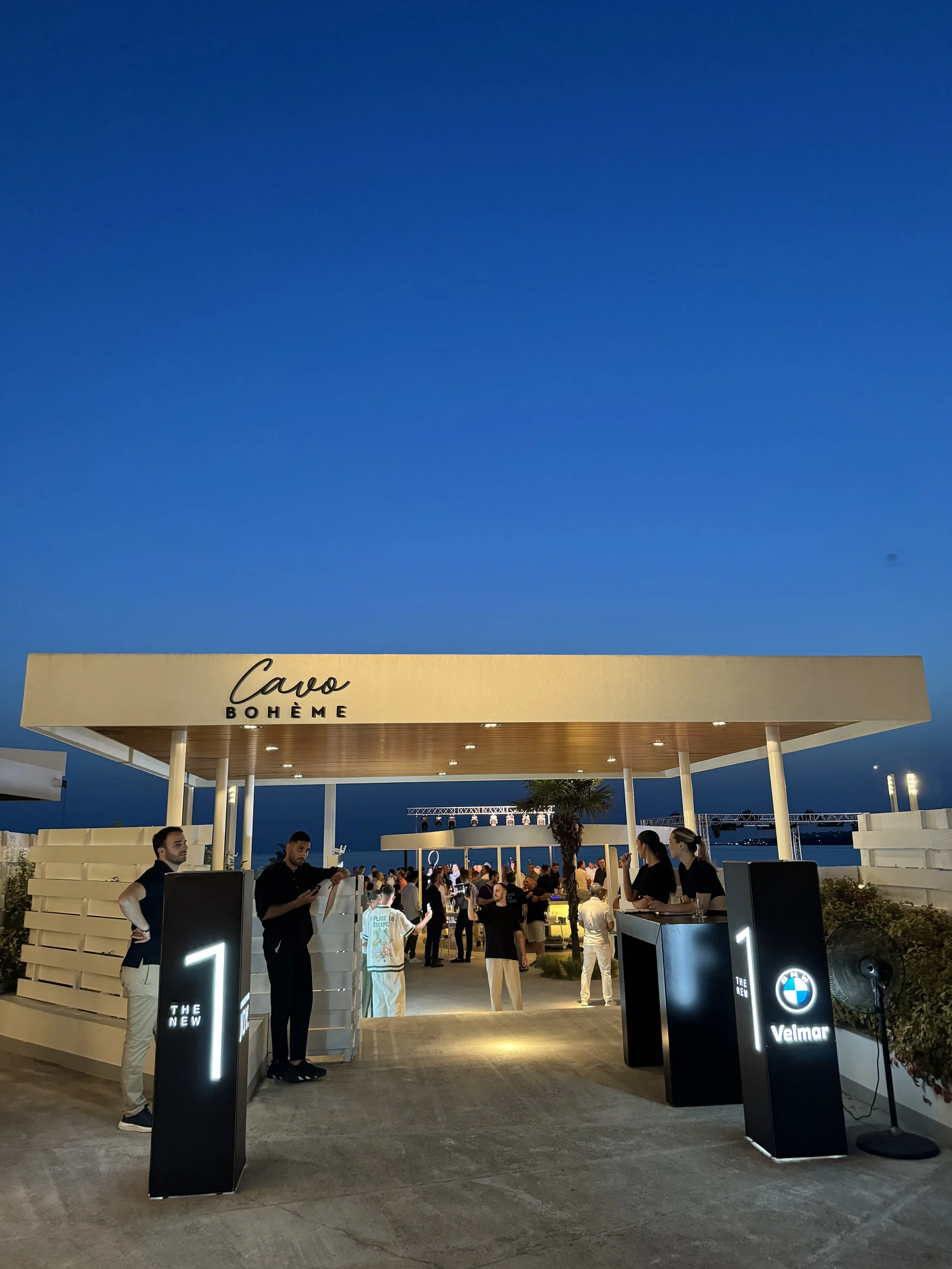 Entrance to a nightclub called Cavò Bohème with people gathering, illuminated signs, and a clear evening sky.