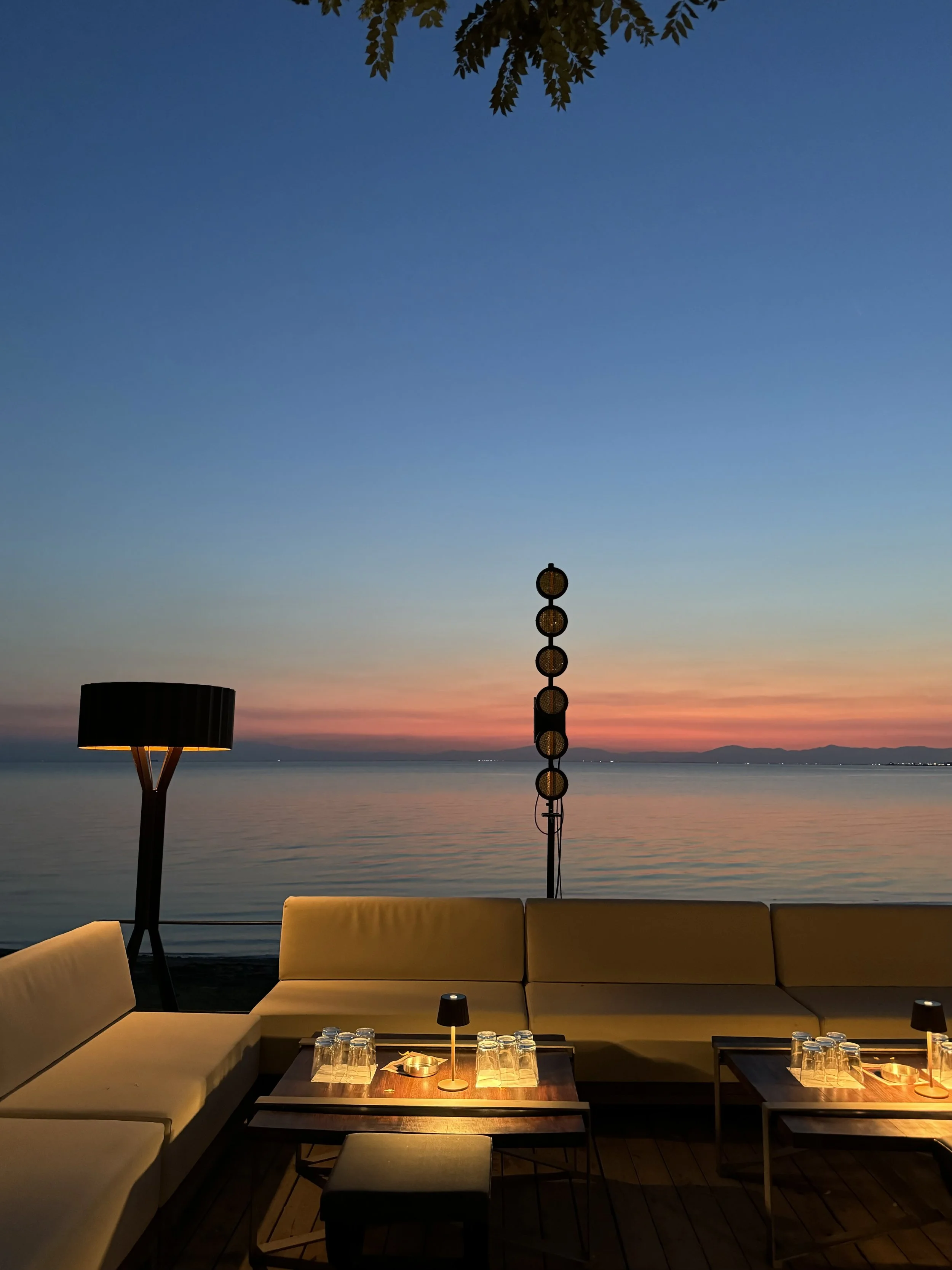Outdoor seating area with beige couches, small tables with glassware and tables lamps, overlooking a calm body of water at sunset with colorful sky, distant mountains, and leaves hanging overhead.