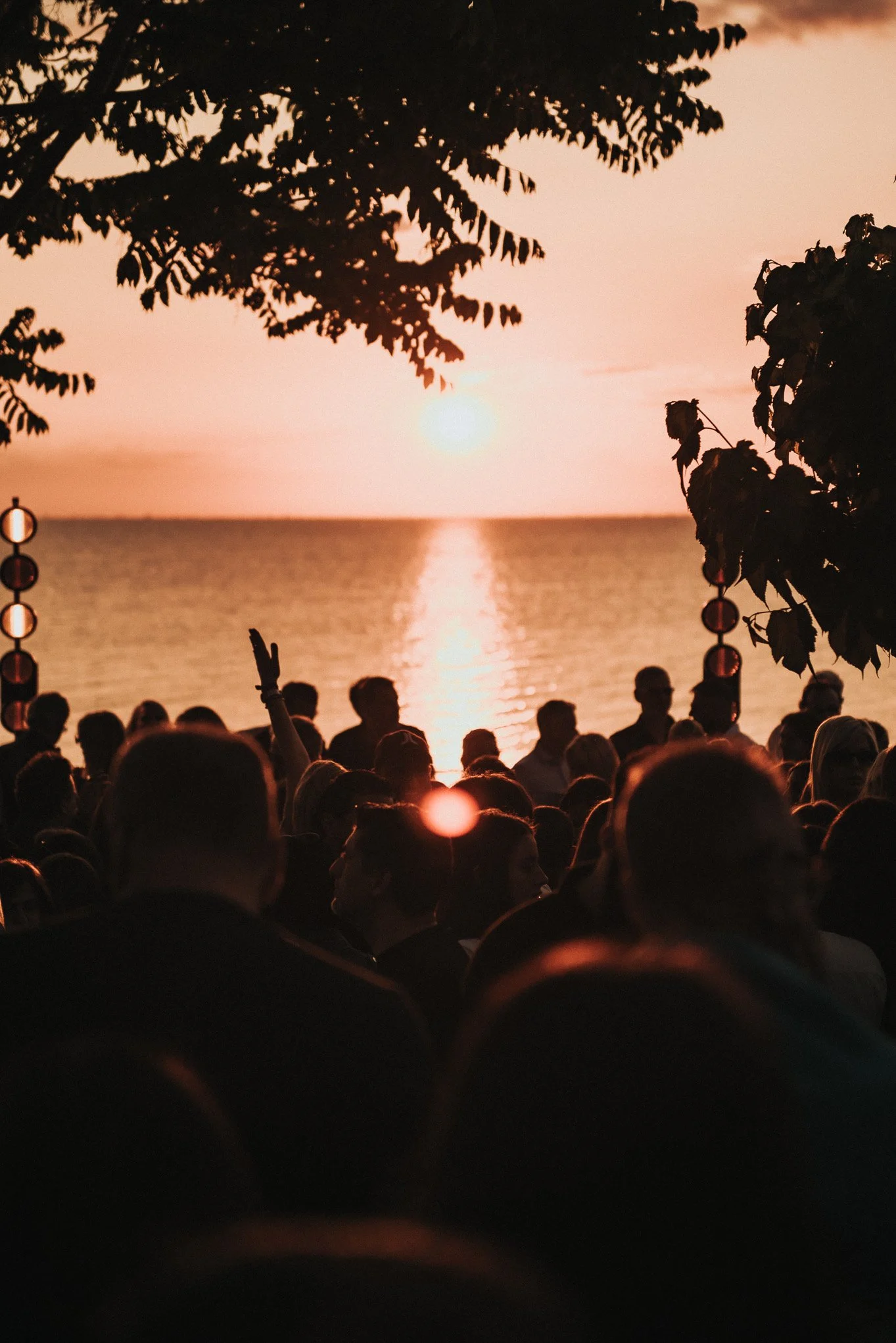 A large crowd of people gathered outdoors during sunset by a body of water, with some raising their hands. The scene is framed by trees on either side, with the sun near the horizon casting an orange glow over the water and silhouettes of the crowd.