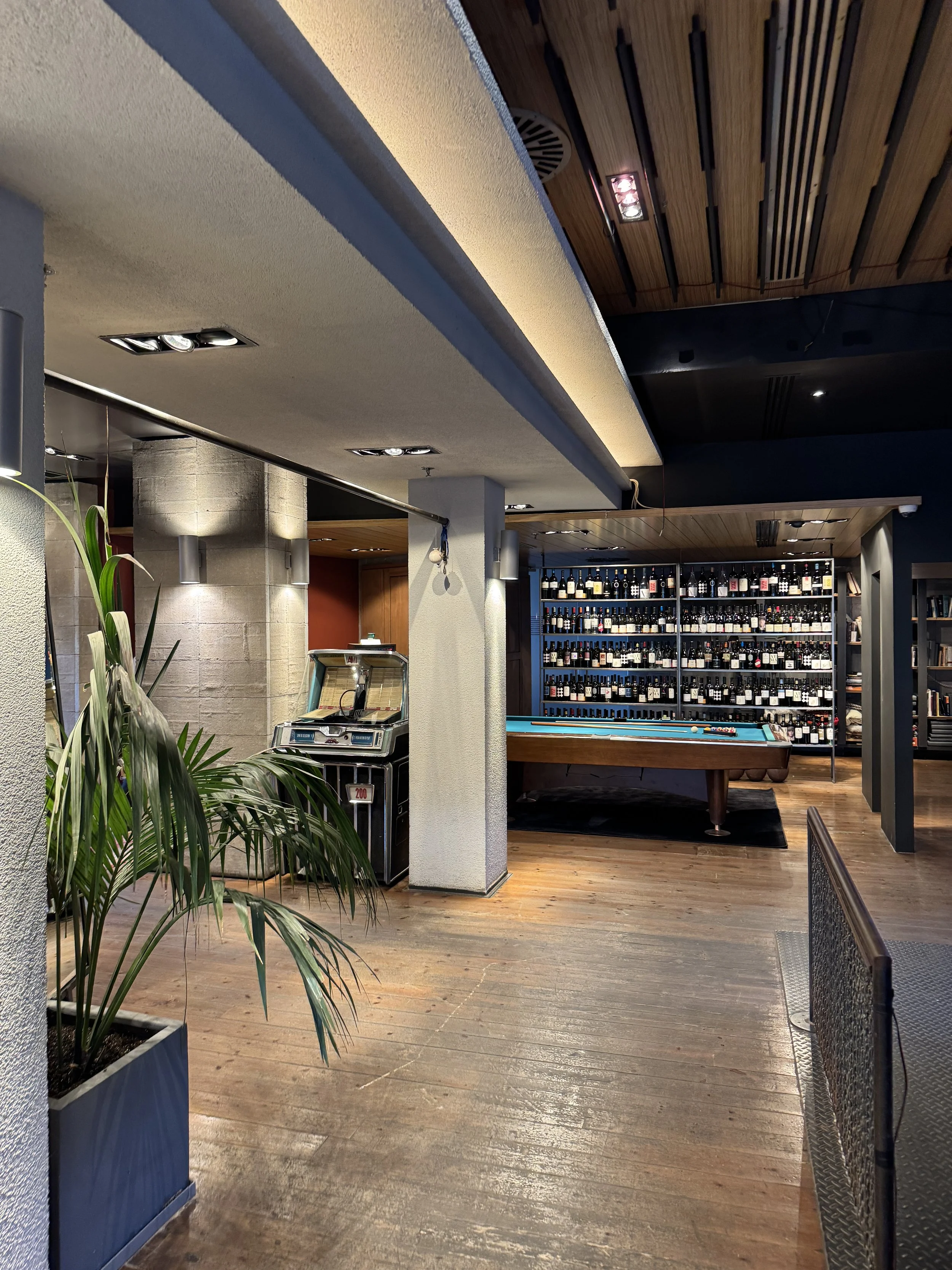 Interior of a stylish bar with a pool table, a large wine rack, and a jukebox, featuring wooden and modern decor.