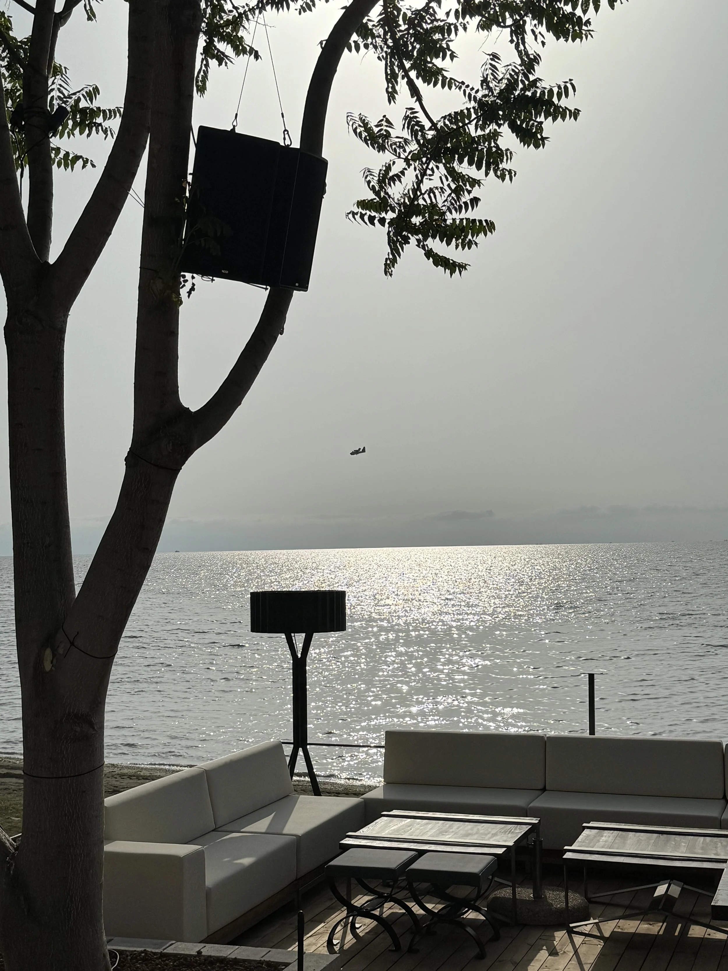 Outdoor seating area with white couches and tables overlooking a body of water, with a tree in the foreground supporting speakers, and an airplane flying in the sky.