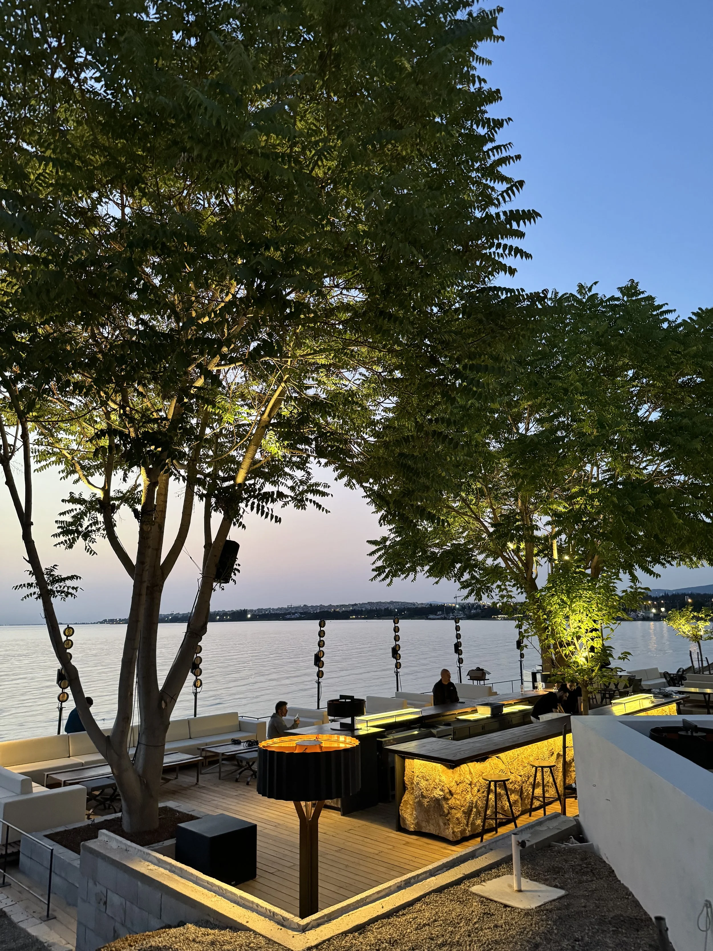 Outdoor lounge area by the water at dusk with trees, seating, and warm lighting.