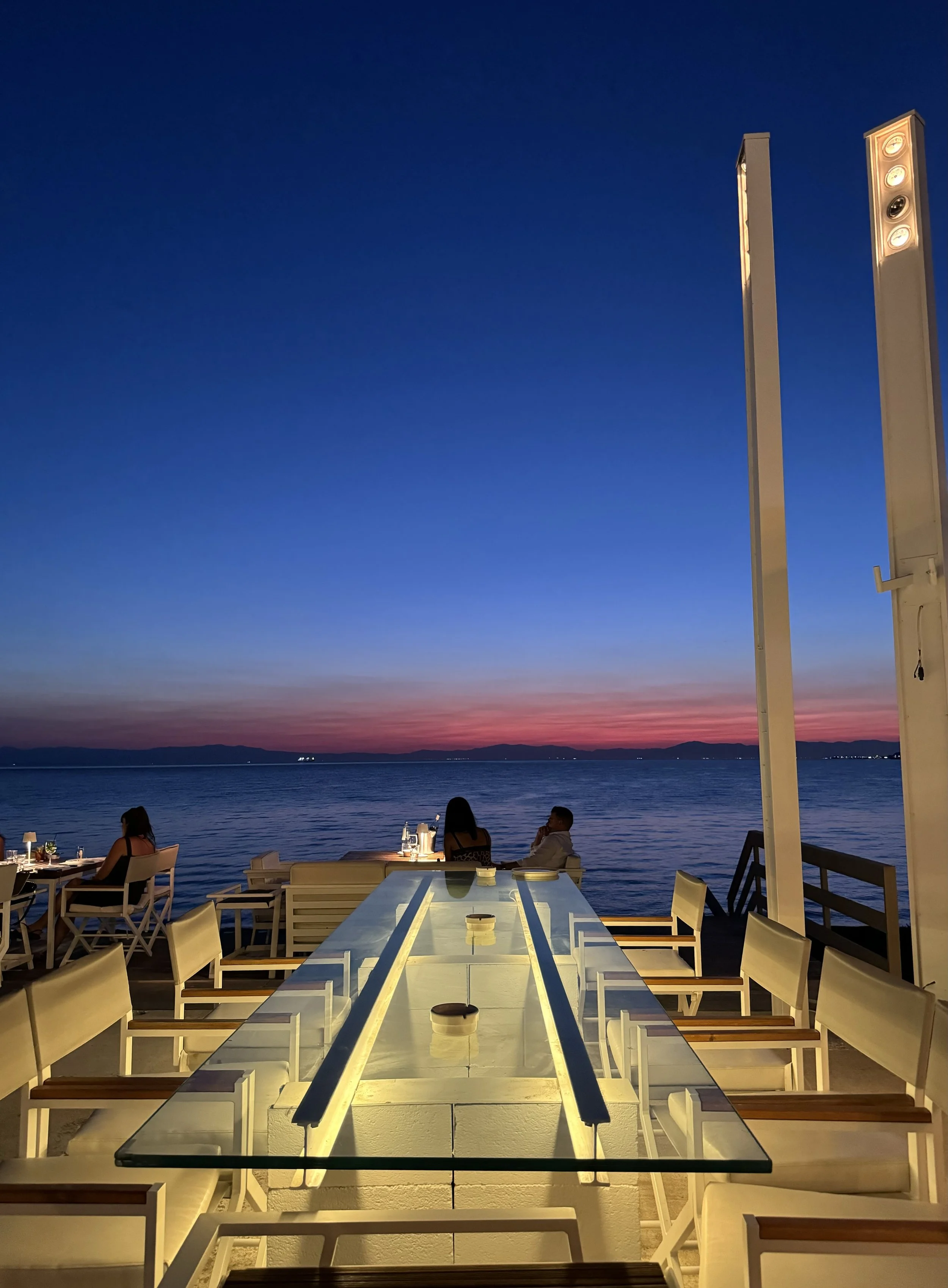 A sunset view from an outdoor dining area overlooking a body of water with distant mountains. The scene includes a long glass table, white chairs, and a few people seated, enjoying the evening.