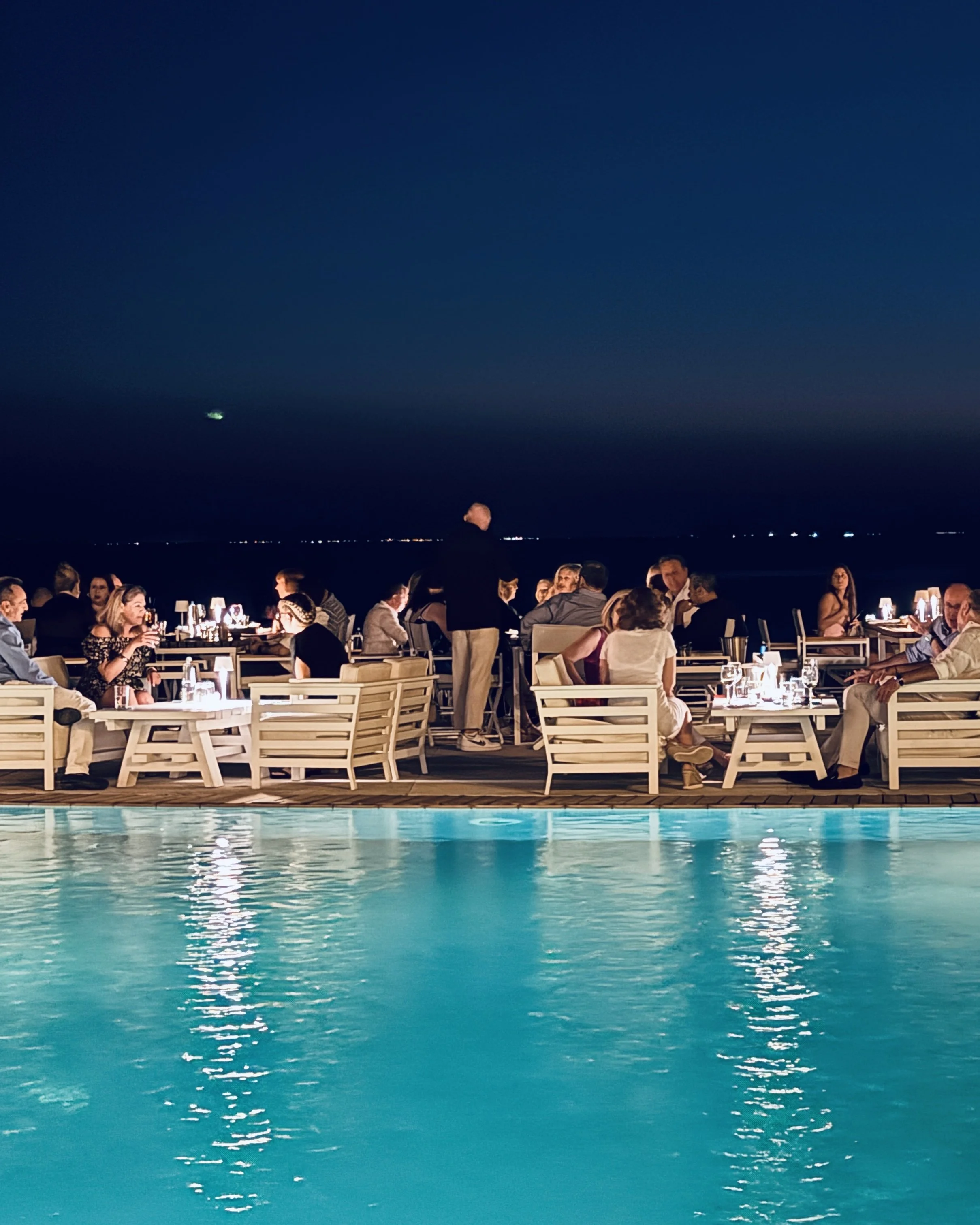 People dining at outdoor restaurant by a pool at night with city lights in the distance.
