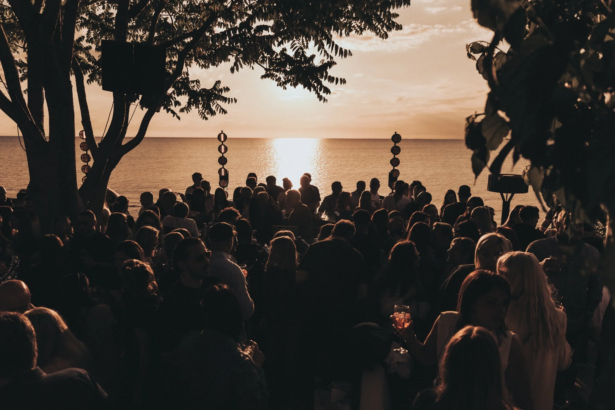 Crowd of people gathered by the seaside during sunset, with trees framing the scene and the ocean reflecting the sunlight in the background.