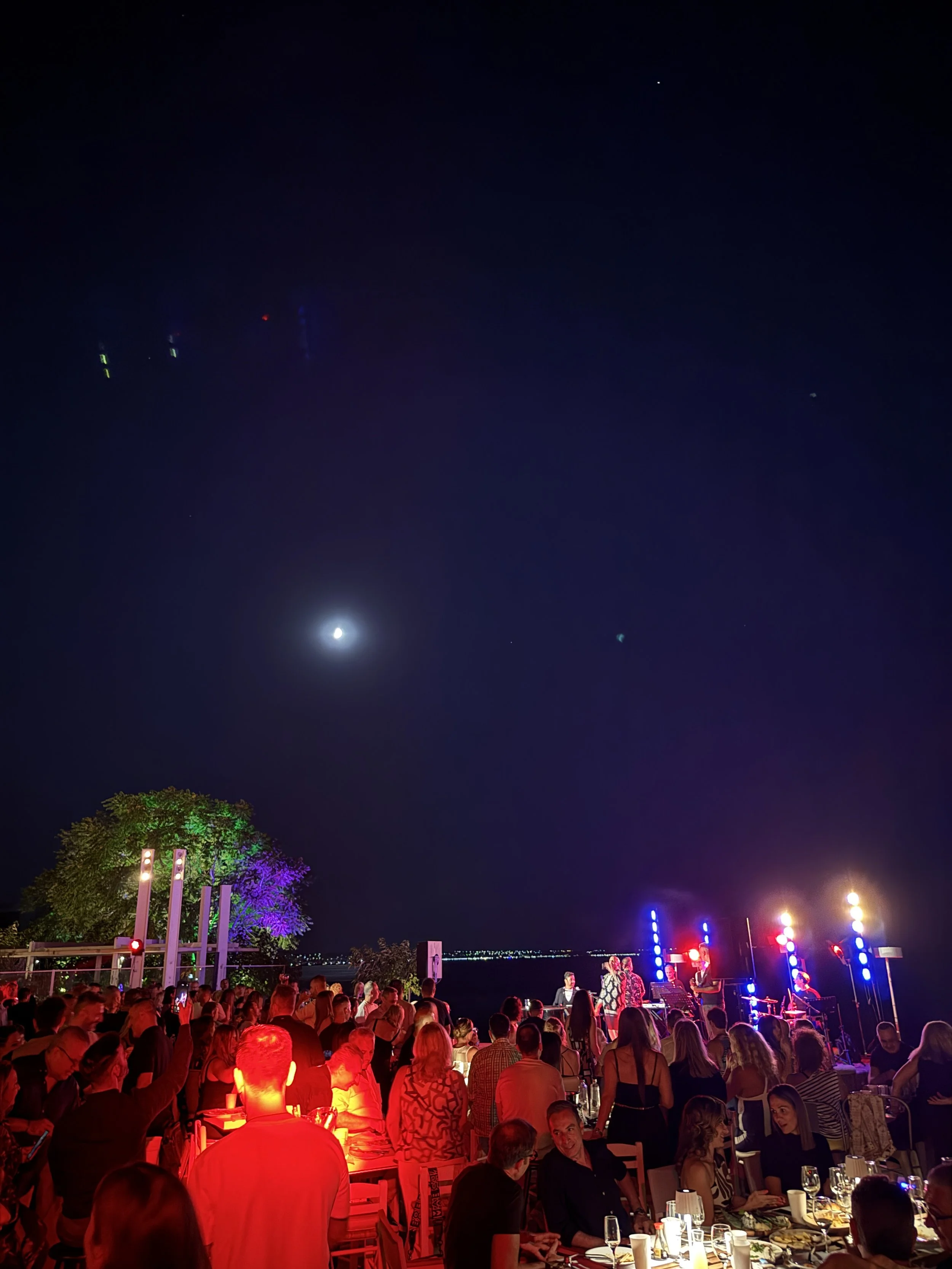 People dining outdoors at night with a view of the moon and city lights, stage with colorful lights, trees, and a dark sky.
