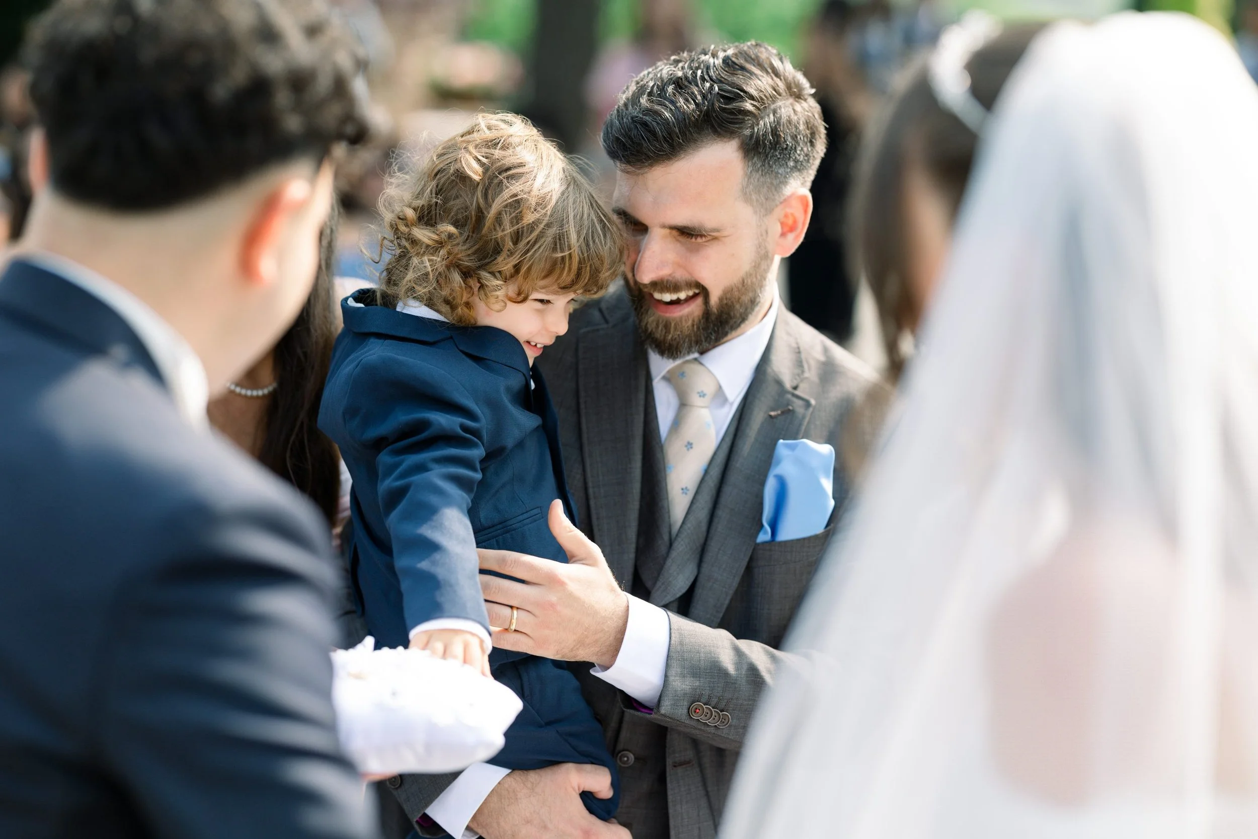 A man with a beard and a suit holding a young boy dressed in a blue suit at what appears to be an outdoor wedding. The man and boy are smiling and leaning their heads close together, with other blurred people in formal attire around them.