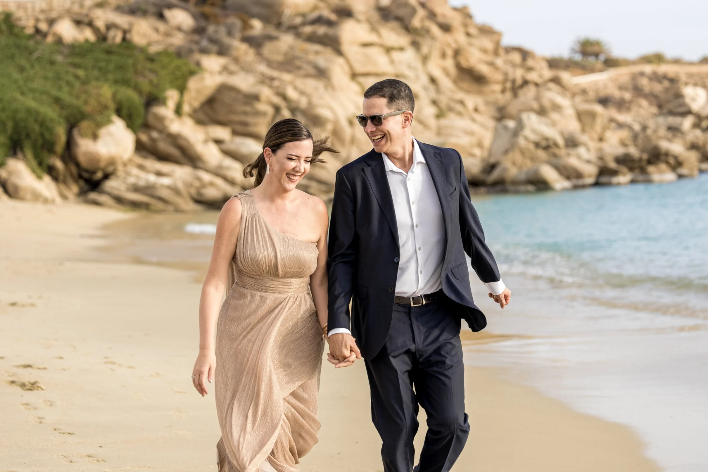 A couple is walking hand in hand along a beach, smiling and enjoying each other's company. The woman is wearing a beige dress, and the man is in a navy suit with sunglasses. There are rocks and a hillside in the background with green vegetation.