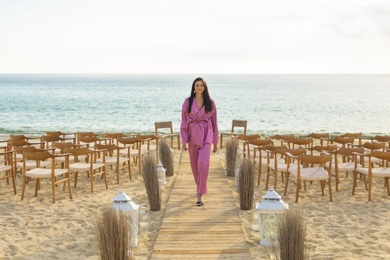 A woman in a pink robe walking down a wooden aisle on a beach, with chairs set up on either side for an outdoor event by the sea.