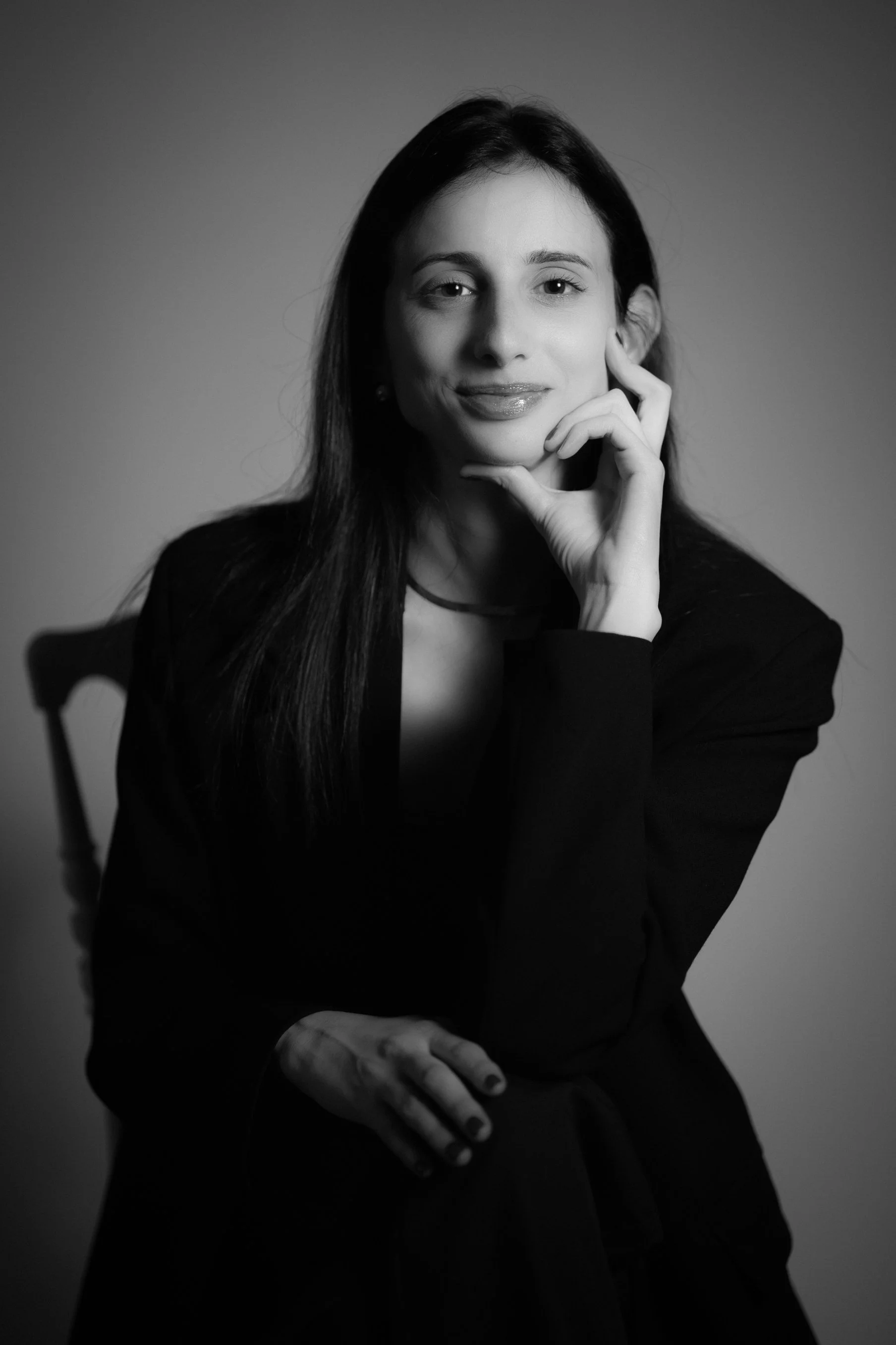 Black and white portrait of a woman with long dark hair, smiling softly, resting her chin on her hand, in a professional setting.
