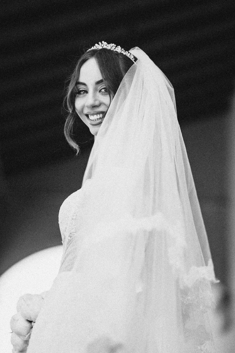 A smiling bride wearing a wedding dress, veil, and tiara, looking over her shoulder.