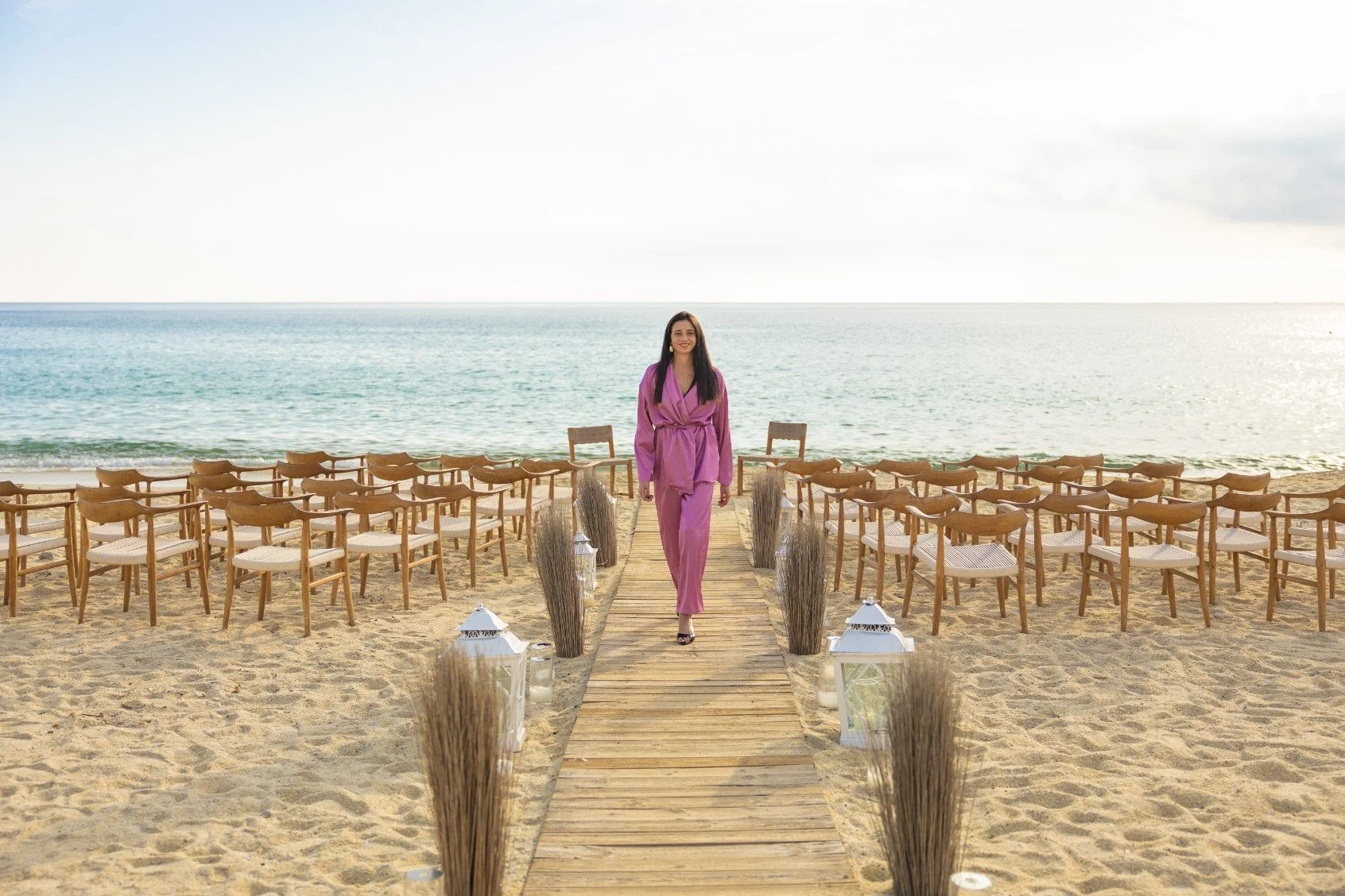 Woman in pink outfit walking down a wooden aisle on the beach with chairs and lanterns, facing the ocean, during daytime.