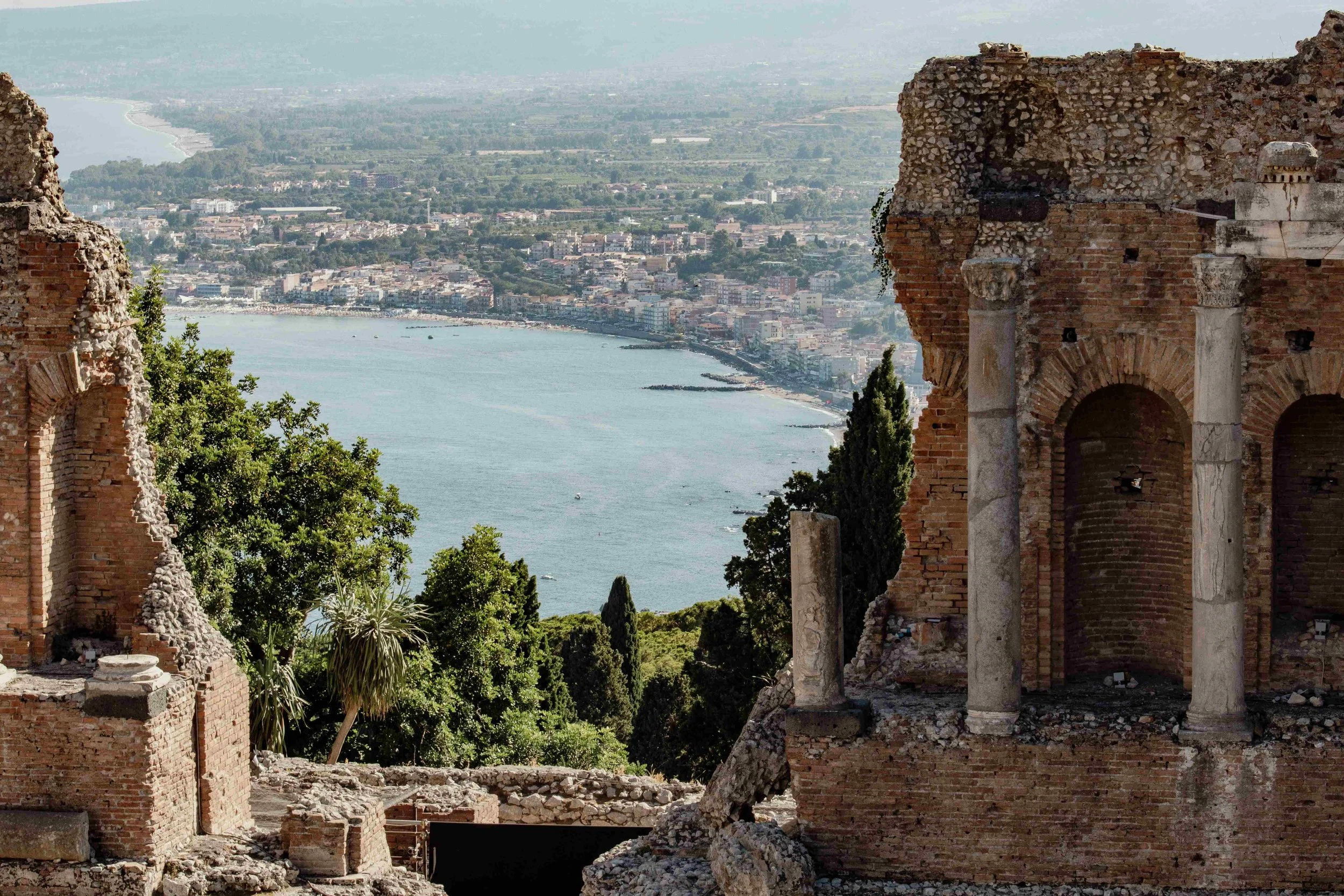 Taormina Ruins of an ancient structure with brick and stone walls overlooking a coastal city with buildings, trees, and a river or lake in the distance.