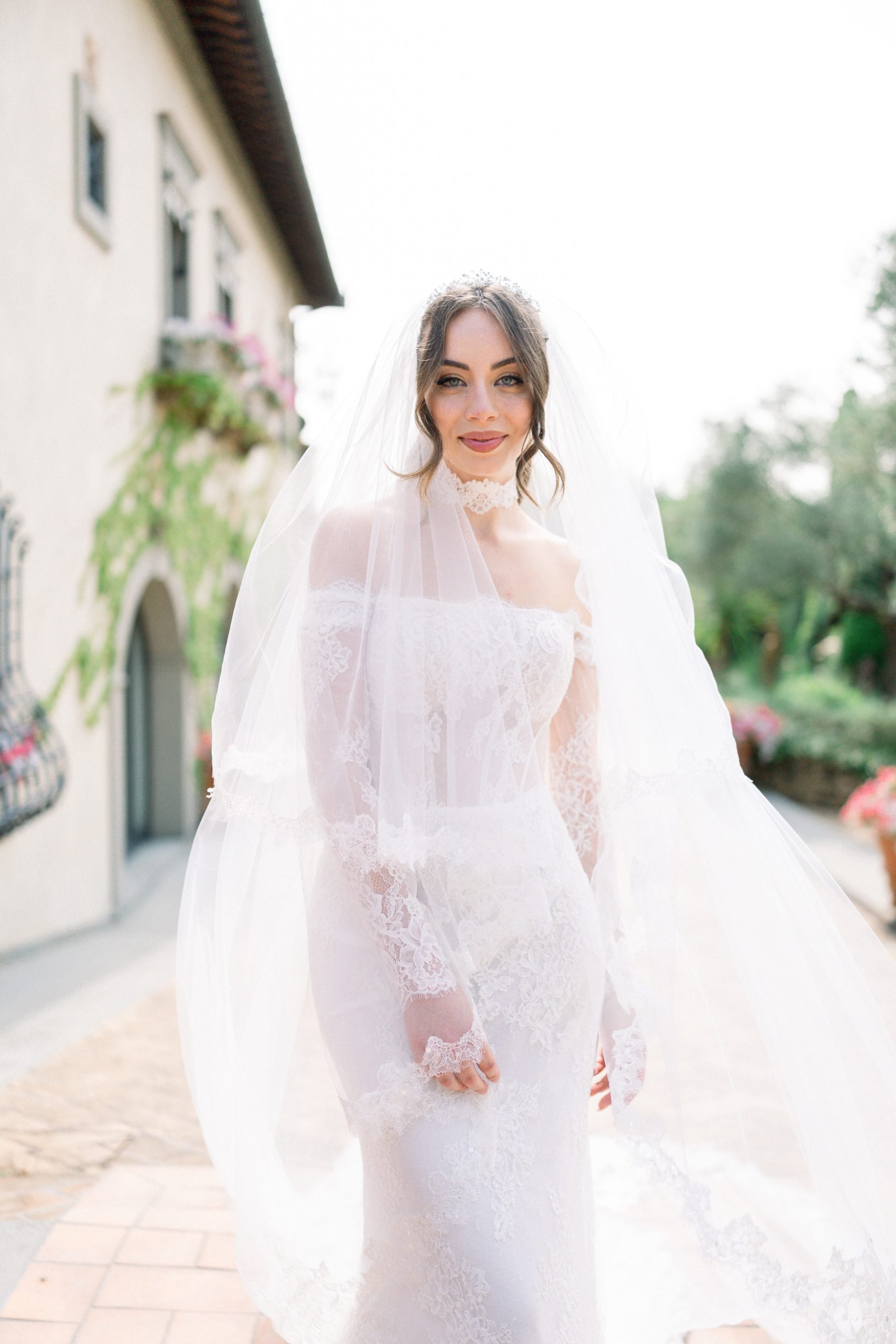 A bride in a white lace wedding gown and veil, standing outdoors next to a building with flower boxes on the windows, smiling softly at the camera.