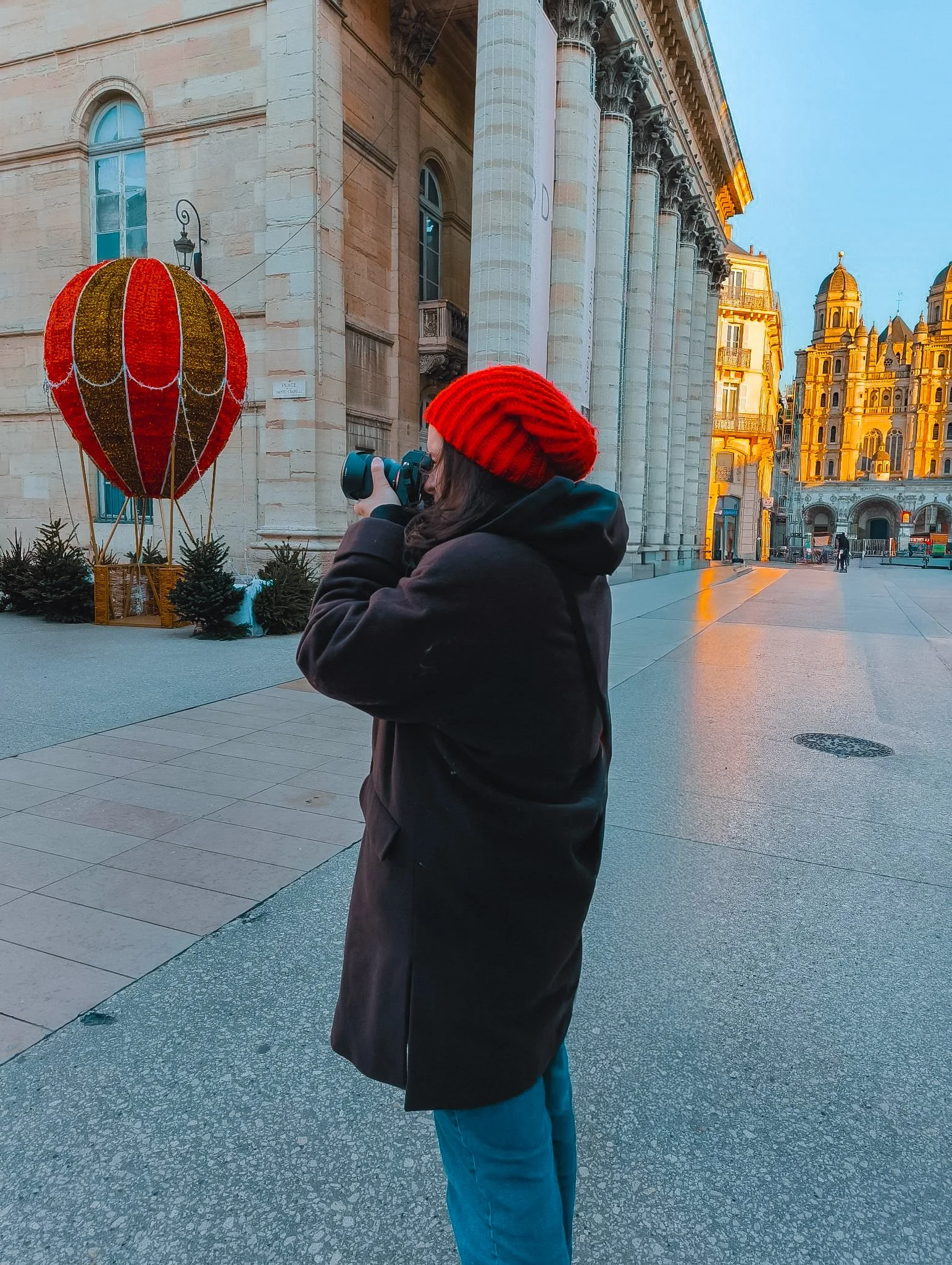 Une femme portant un bonnet rouge et une veste sombre, tenant un appareil photo, se trouve dans une place avec des bâtiments historiques en arrière-plan, décorée avec une lanterne en forme de ballon