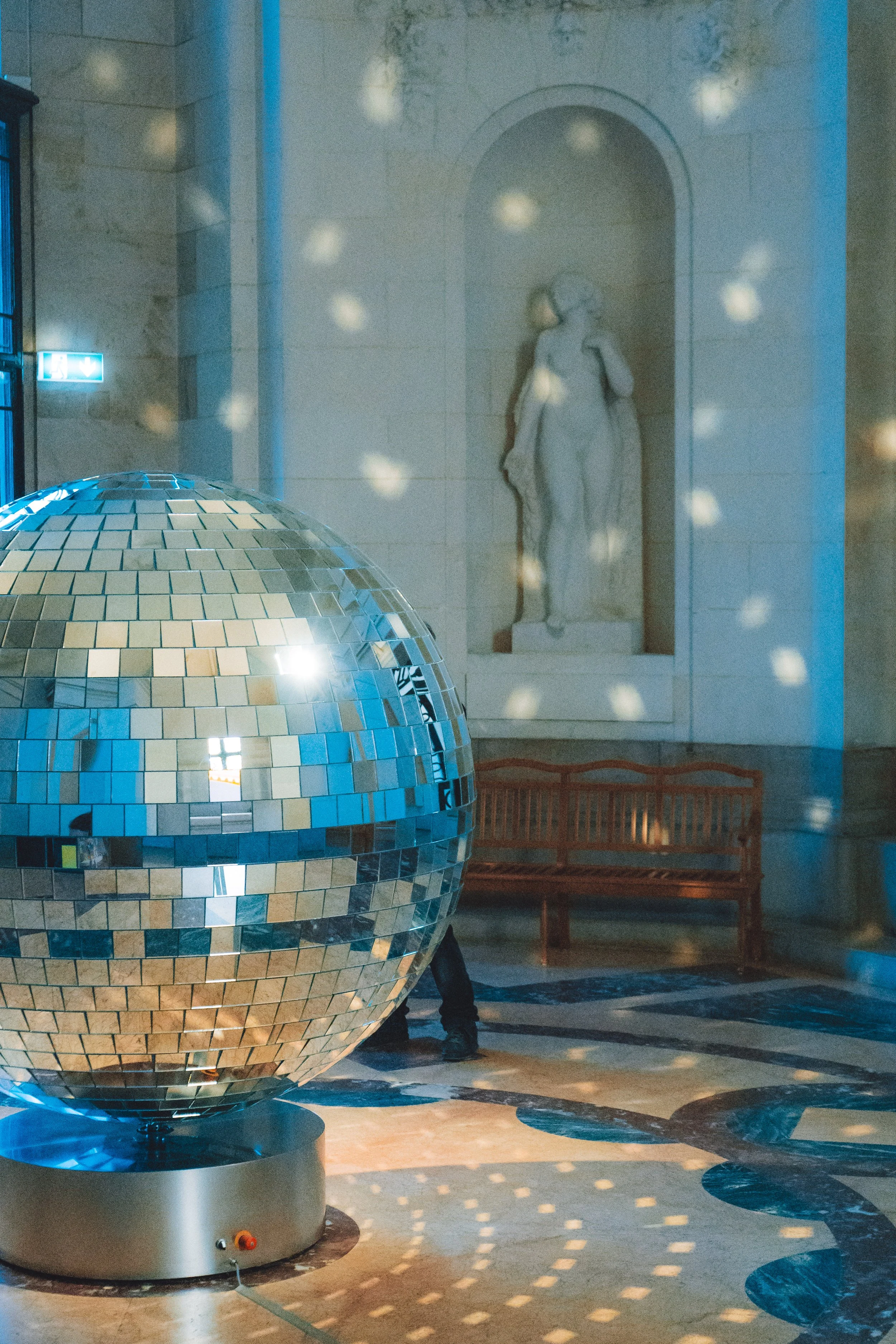 Une sphère disco en miroir reflète la lumière dans un espace intérieur. En arrière-plan, une sculpture en marbre d'une femme debout dans une niche et un banc en bois sont visibles. Des effets de lumière projettent des motifs circulaires au sol.