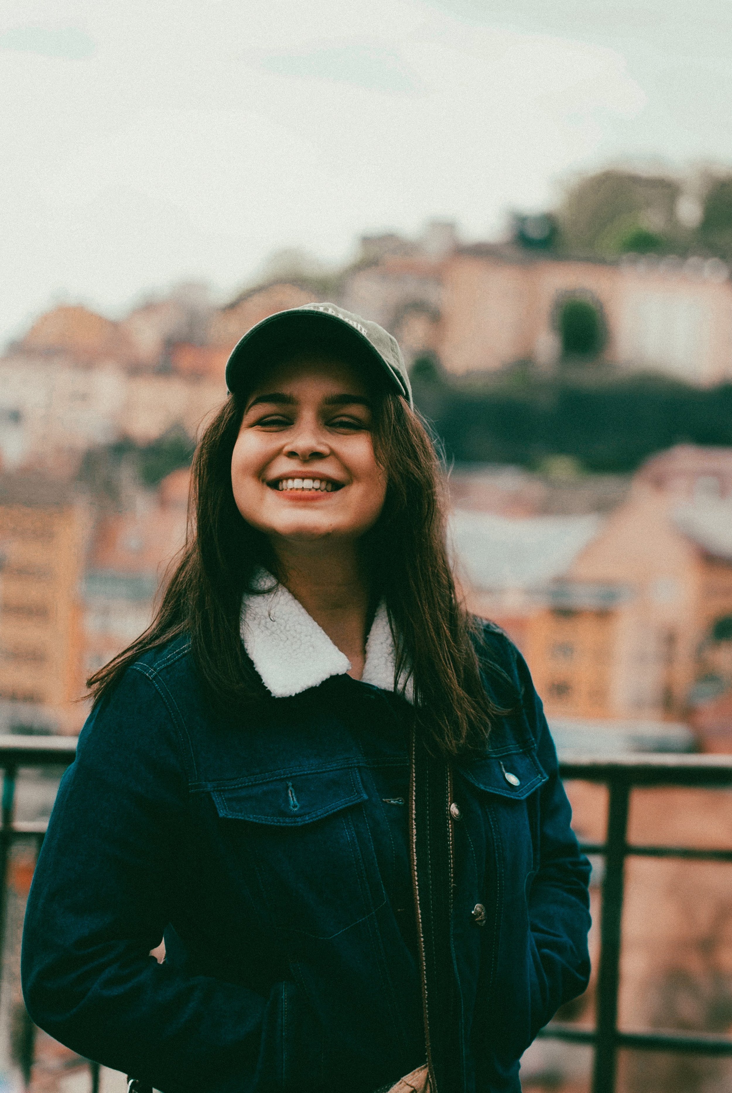 Jeune femme souriante portant une casquette et un blouson en jean, devant un paysage urbain avec des bâtiments en arrière-plan.