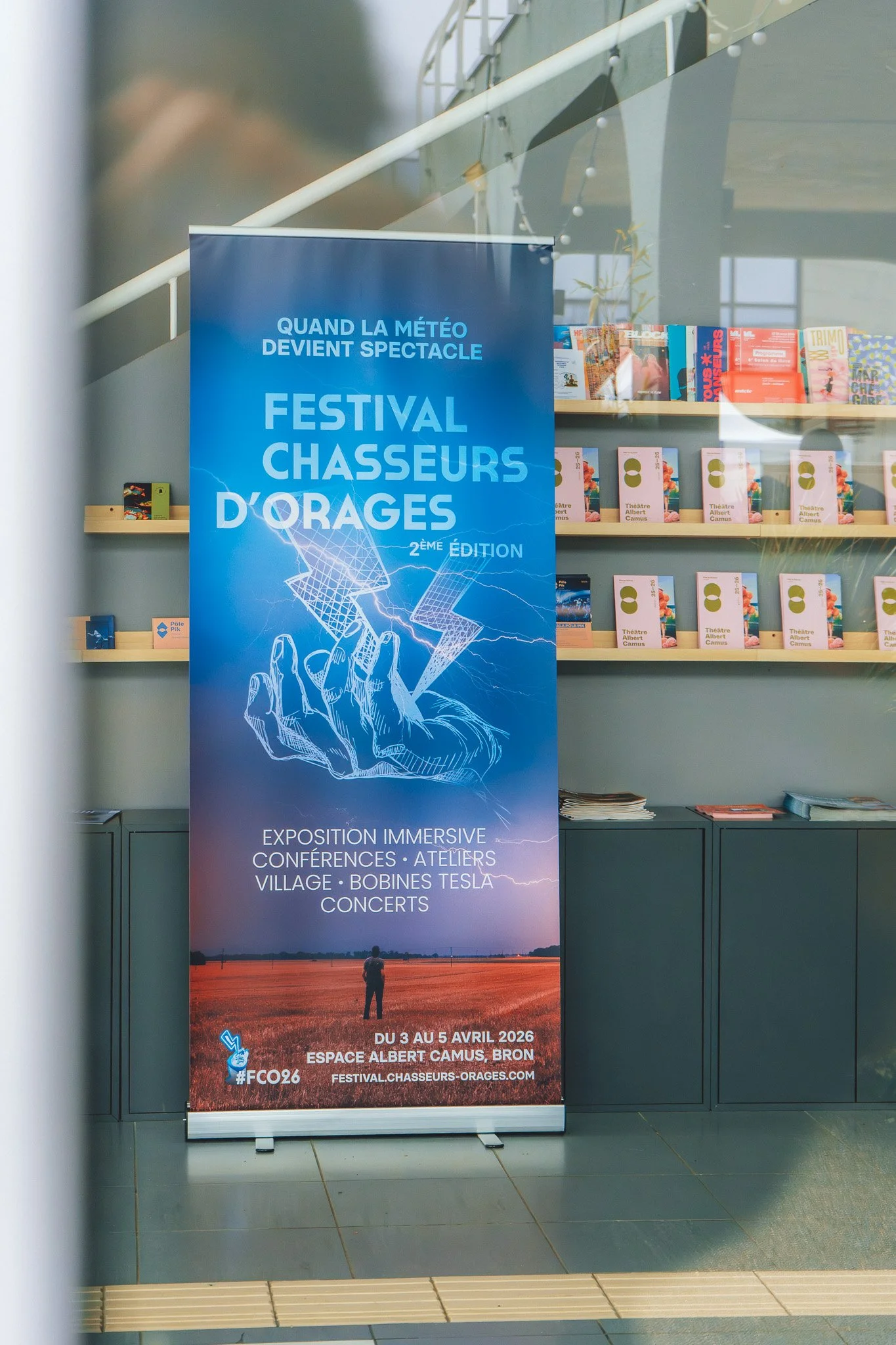 Affiche annonçant le Festival Chasseurs d'Orages, qui se déroulera du 3 au 5 avril 2026 à l'Espace Albert Camus à Bron, avec un fond d'un paysage de champs et un ciel orageux, et des détails sur les conférences, ateliers, village, bobines Tesla et co