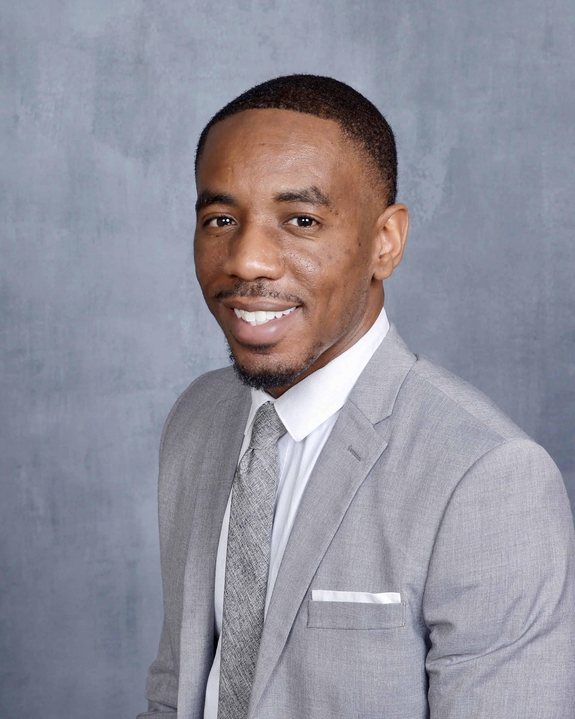 A man wearing a grey suit, white shirt, and grey tie, smiling at the camera with a grey background.