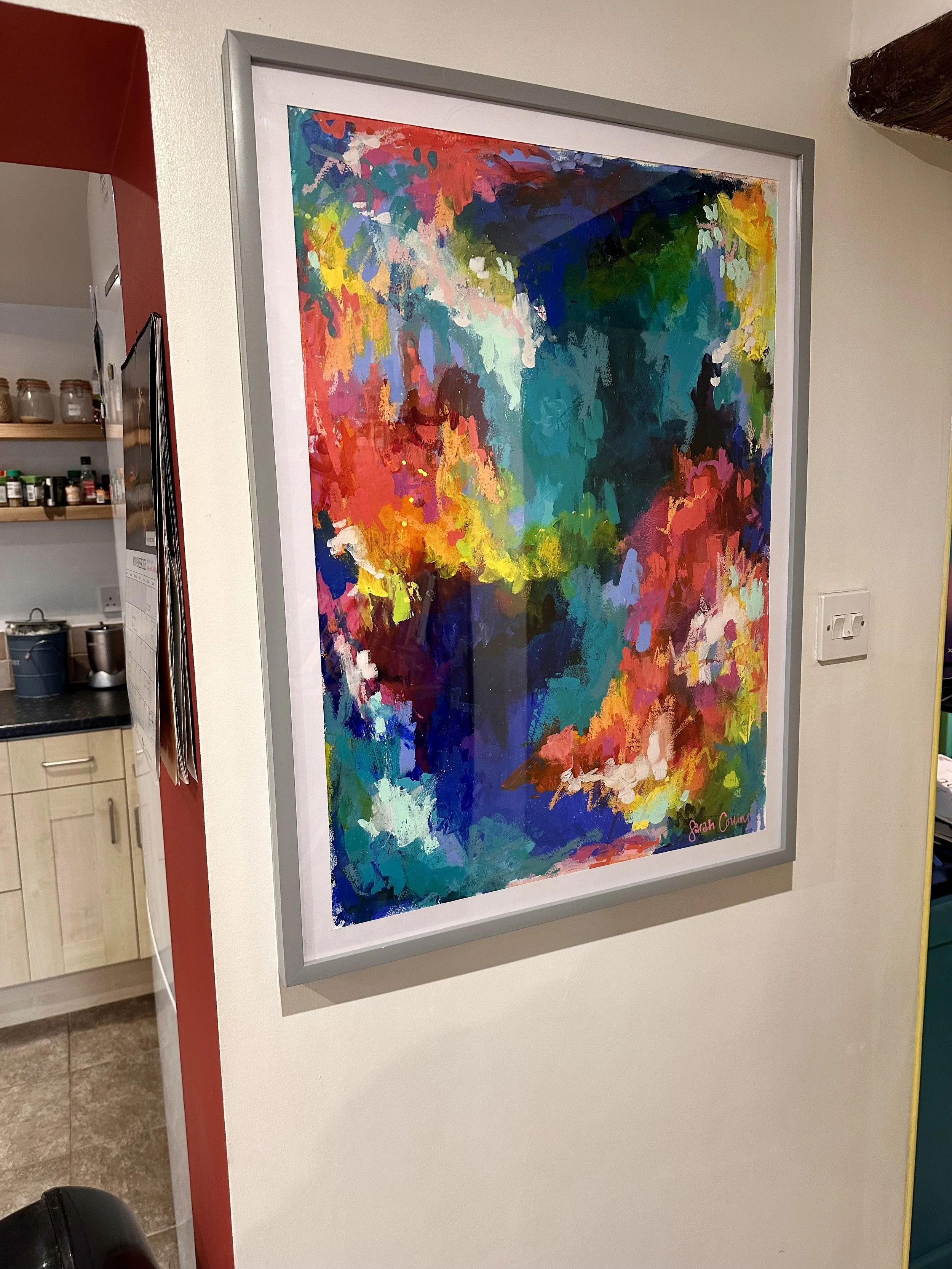 An abstract, colorful painting with vibrant brushstrokes in reds, blues, greens, yellows, and pinks, framed and hung on a white wall, signed 'Sarah Cowen' in the bottom right corner.
