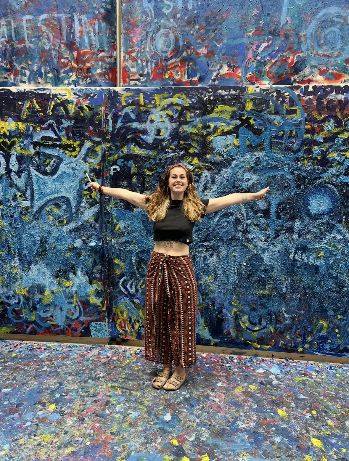 A woman with curly hair and tattoos on her stomach standing with arms wide open in front of a large, colorful abstract painting. The scene appears to be in an art studio or gallery with a paint-splattered floor.